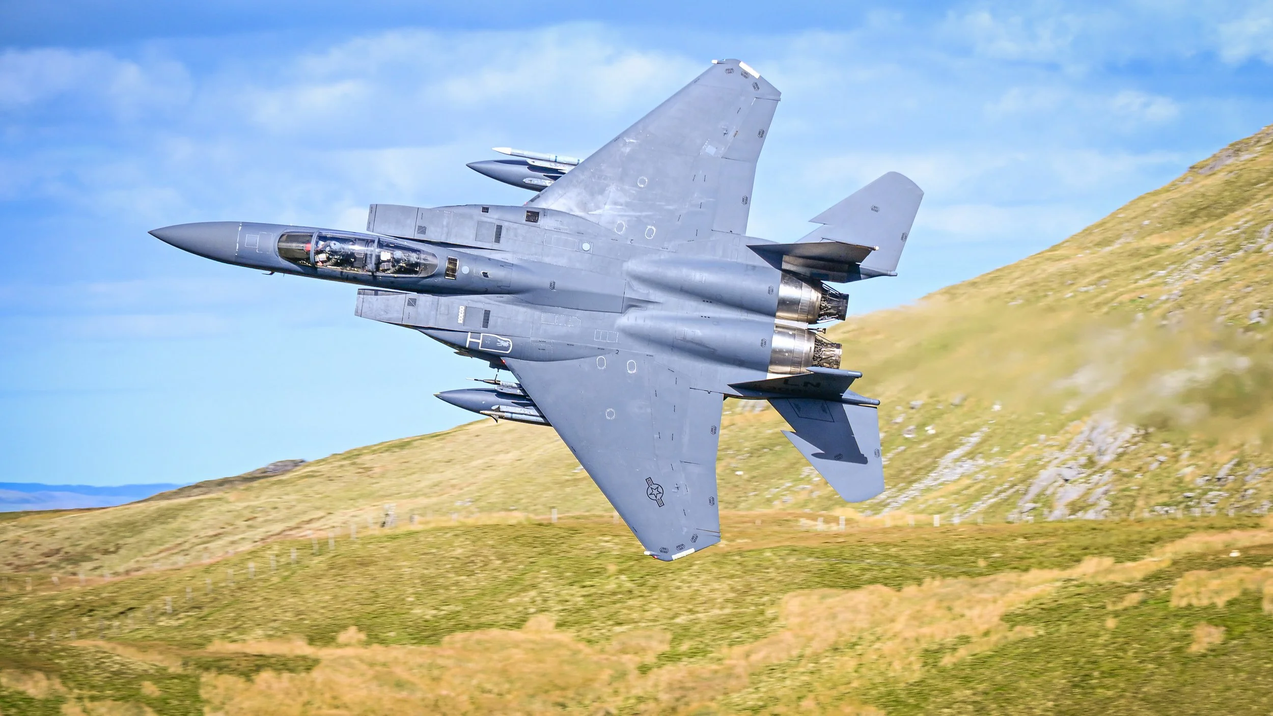 A military fighter jet flying over grassy hills and blue sky.