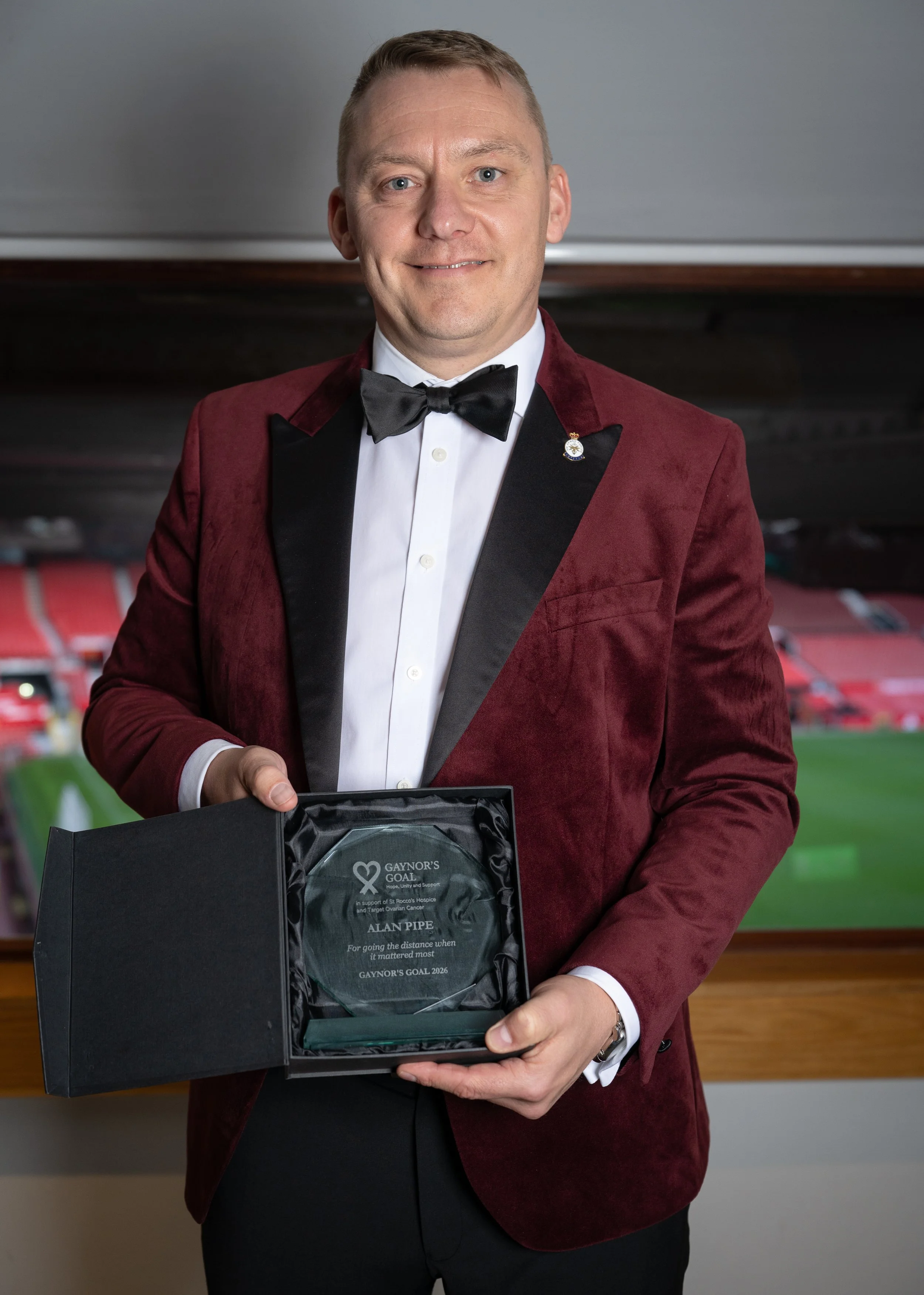 A man in a tuxedo and maroon velvet blazer holding a glass award. The award's engraving mentions gaynor's goal, Alan Pipe, and support for ovarian cancer.