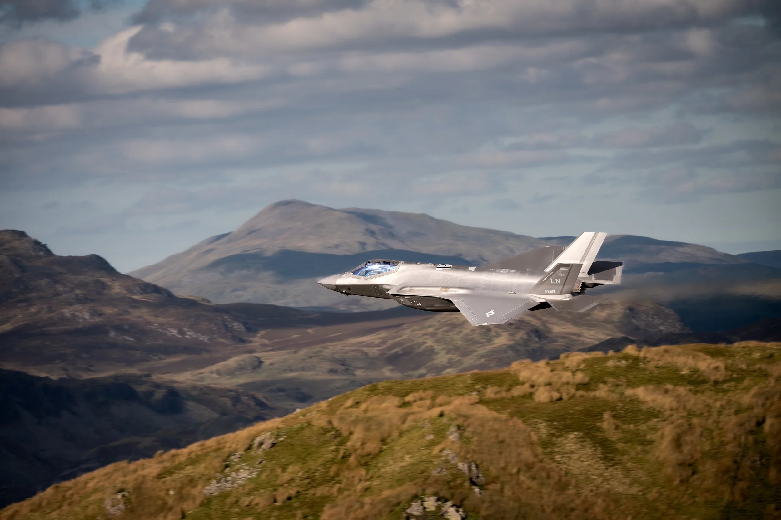 A fighter jet flying over green hills and mountains with a cloudy sky in the background.