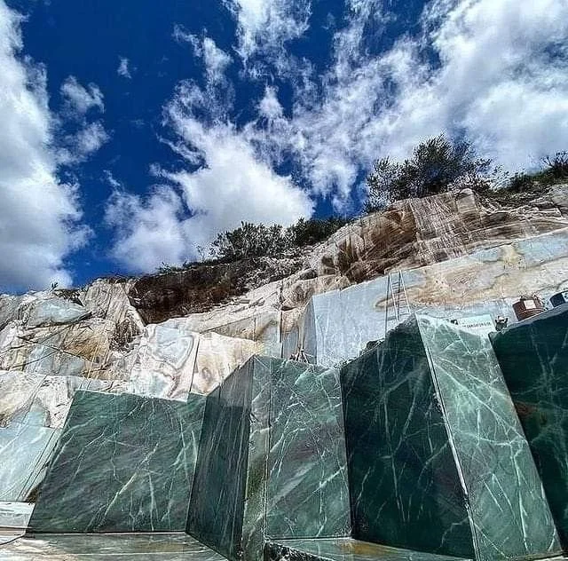 A quarry with large, polished green marble blocks stacked against a rocky cliff under a bright blue sky with scattered clouds.