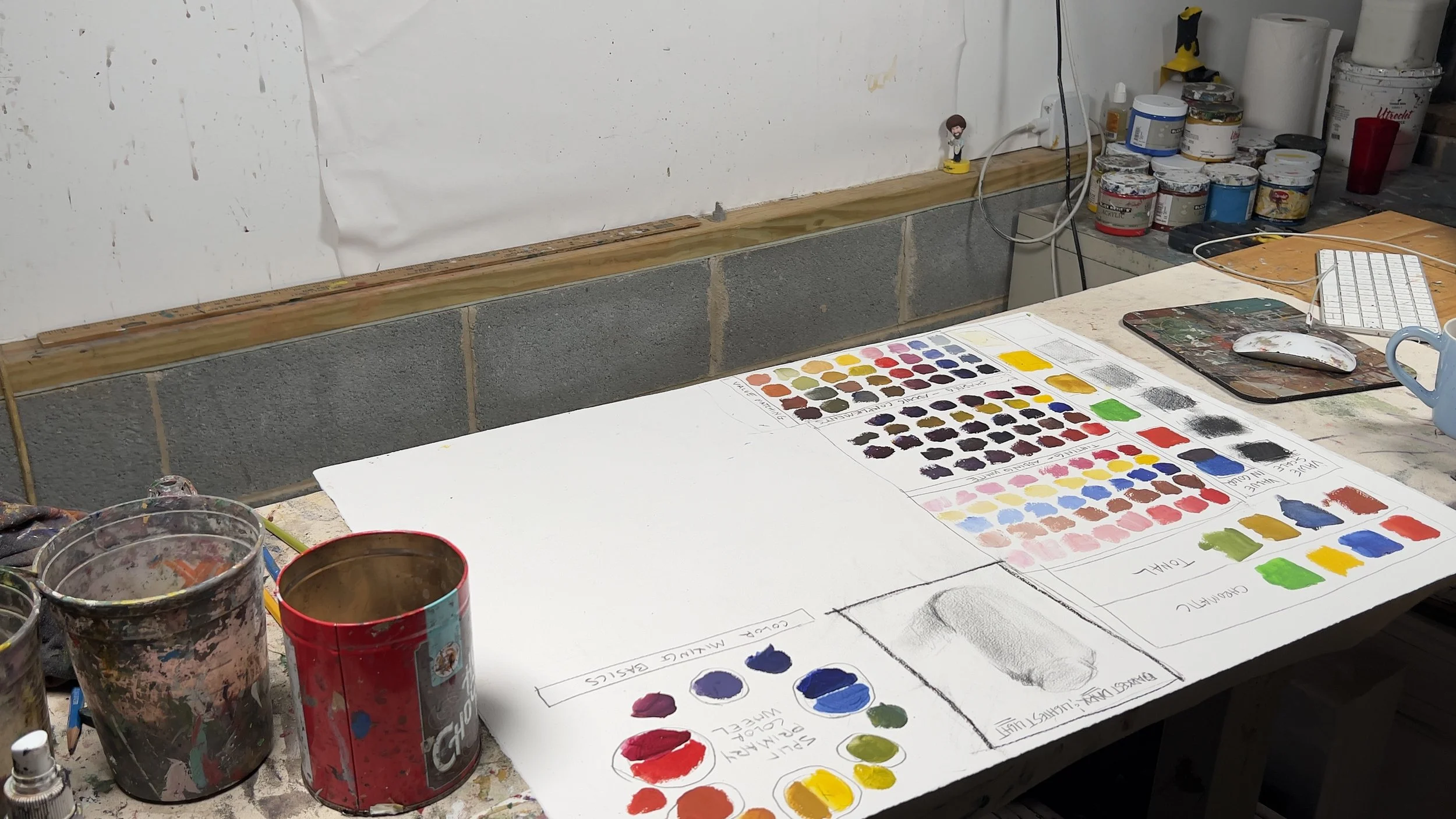 An art studio workspace with paint cans, a color chart, and a worktable, featuring a strip of wooden molding along the wall and various art supplies in the background.