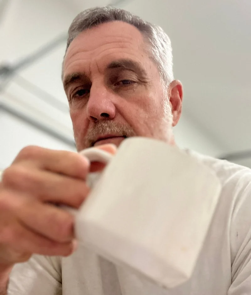 A man with gray hair and a beard drinking from a white mug.