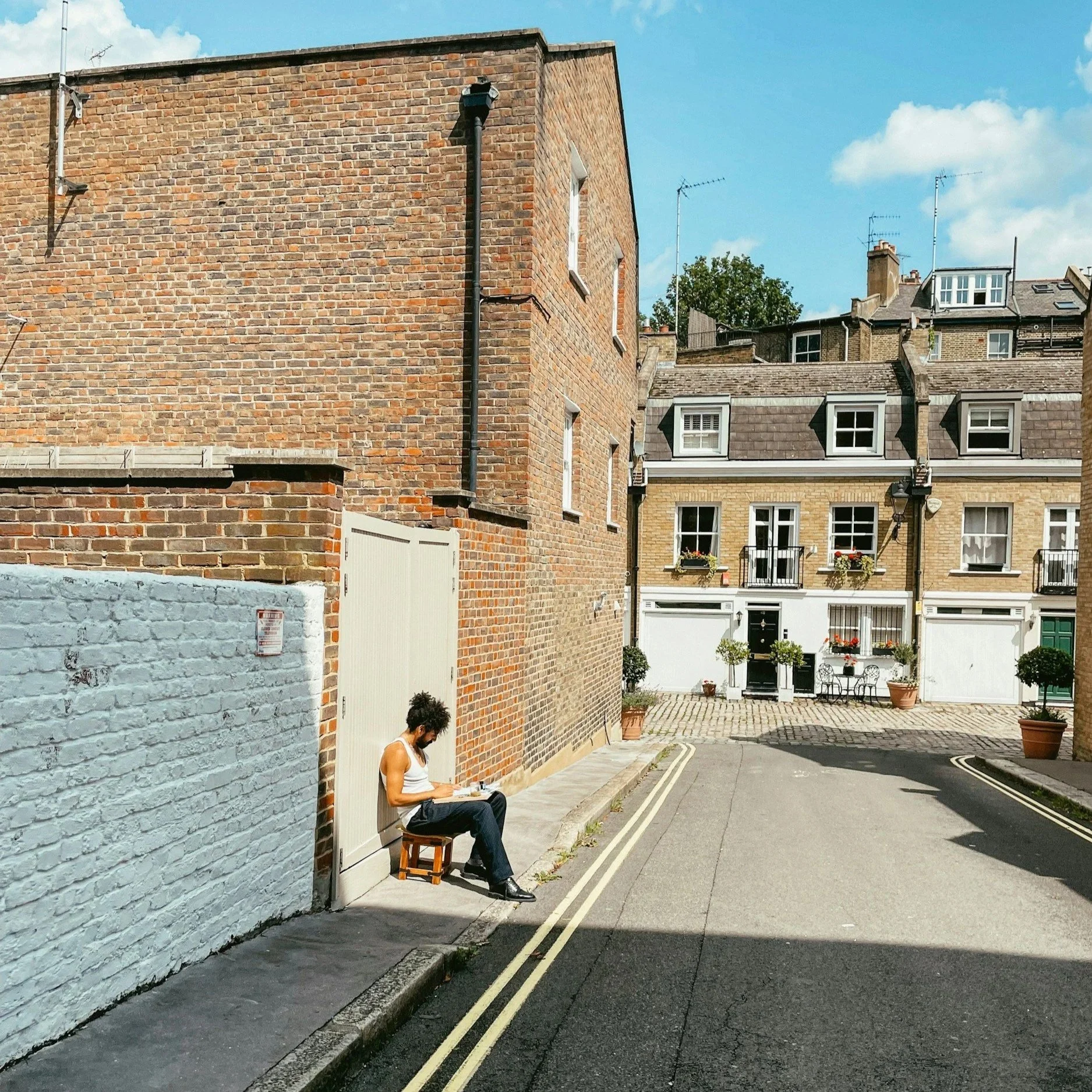 A person sitting on a small wooden stool on a city sidewalk, reading a book, with brick buildings and potted plants in the background under a blue sky.