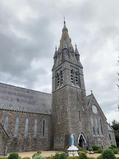 A large stone church with a tall steeple, gothic windows, and a statue of the Virgin Mary at the entrance, under a cloudy sky.