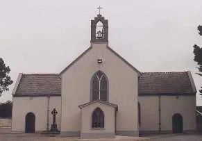 Small white church with a cross on top, a bell tower, and an arched doorway, surrounded by trees.