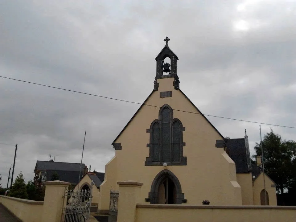 A small church with beige walls, dark gray window trims, and a bell tower topped with a cross under a cloudy sky.