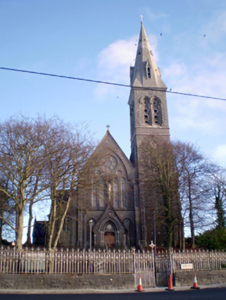A historic church with a tall steeple, stone facade, and arched windows, surrounded by leafless trees and a metal fence, under a blue sky.