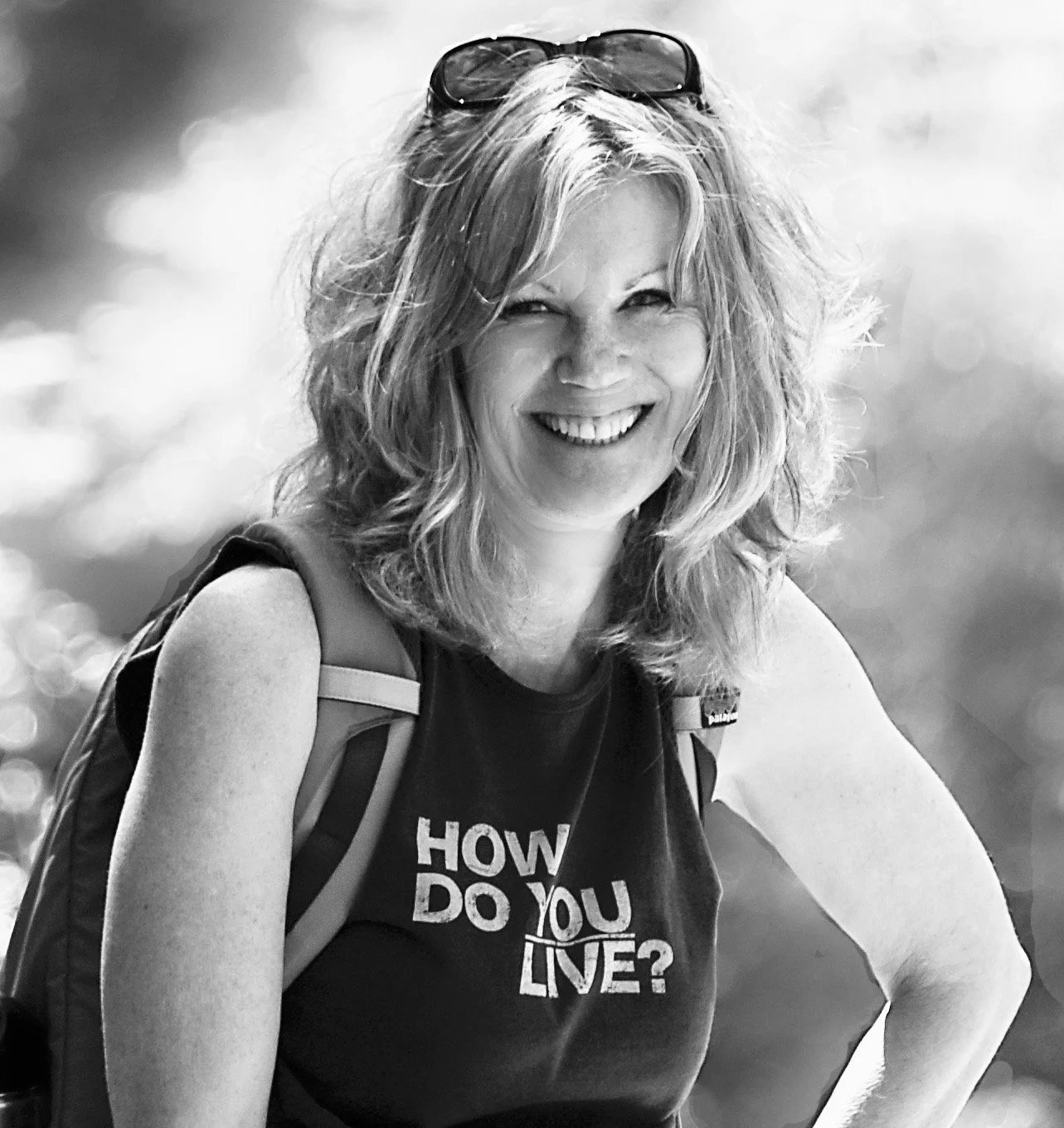 A smiling woman with wavy hair wearing sunglasses on her head, a backpack, and a sleeveless top with the words "How do you live?" printed on it.