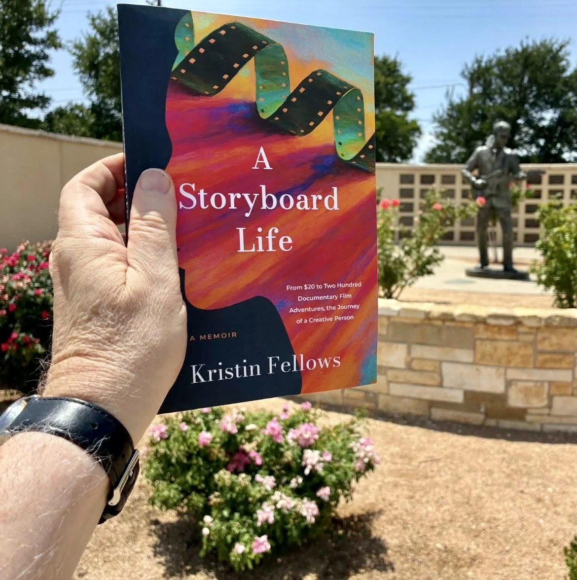 Hand holding a colorful book titled 'A Storyboard Life' by Kristin Fellows in front of a statue of a person with a guitar, surrounded by pink flowers and a brick wall, outdoors on a sunny day.