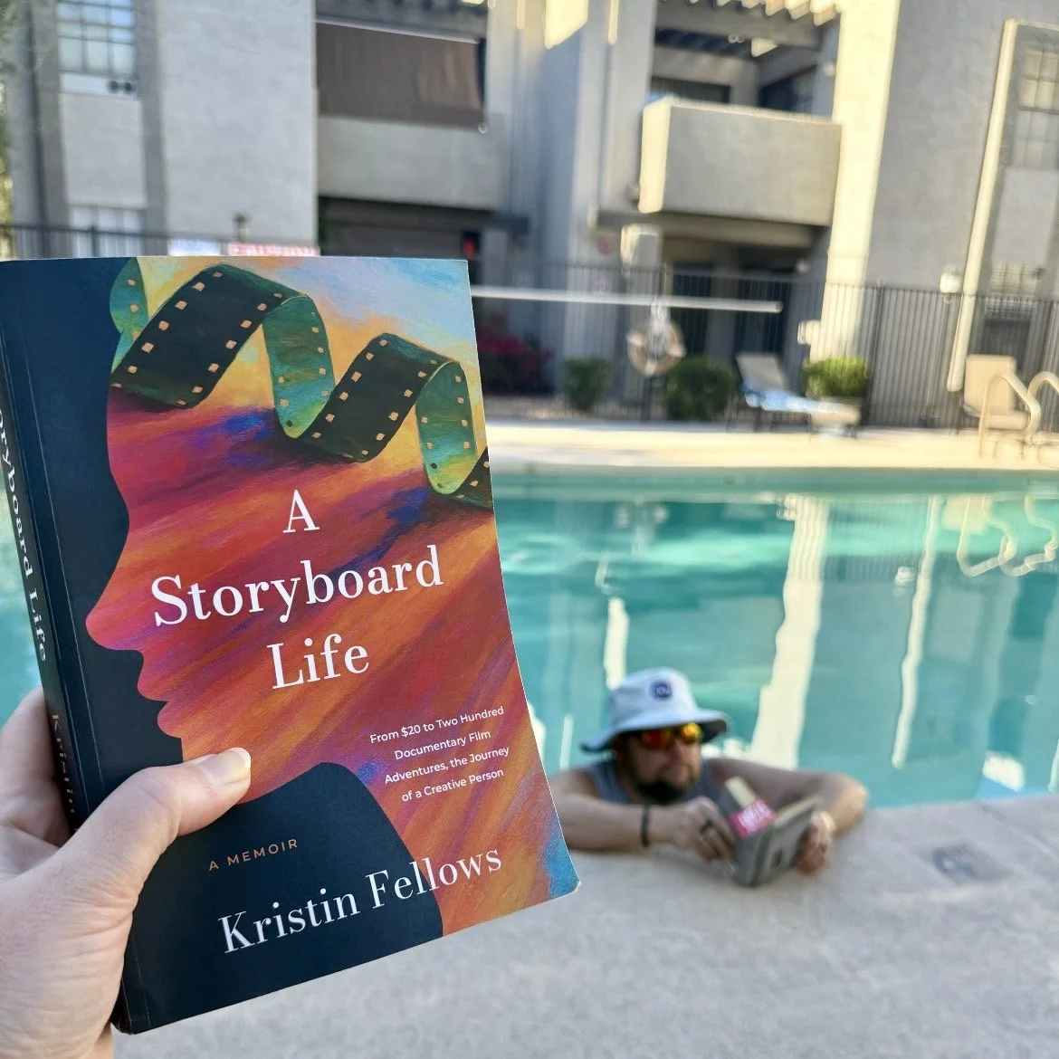 A person holding a book titled 'A Storyboard Life' by Kristin Fellows by a swimming pool, with a man lounging in the pool in the background wearing sunglasses and a white hat.
