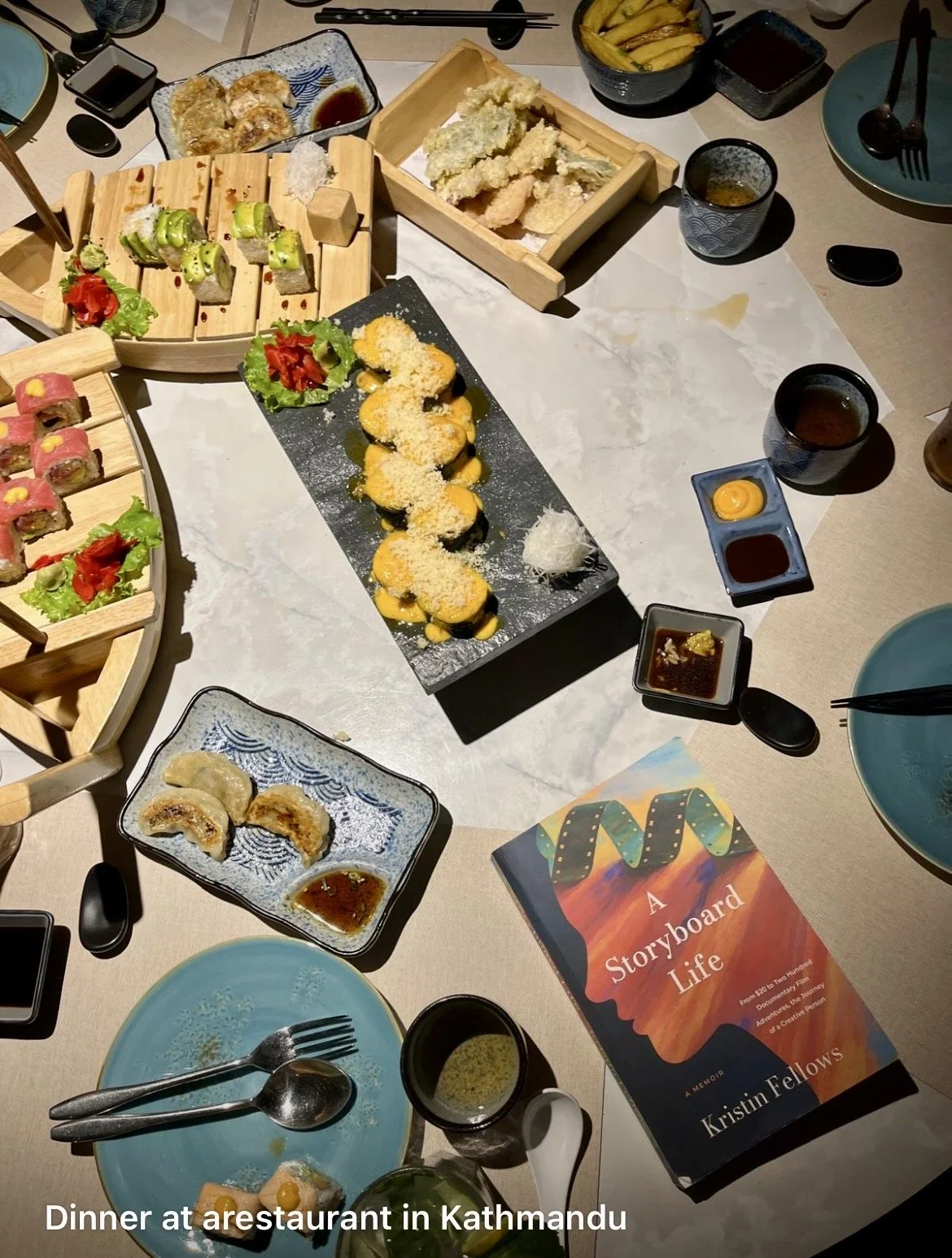 A table filled with various Japanese dishes including sushi rolls, tempura, gyoza, and dipping sauces. There are small cups of tea or soy sauce, plates, chopsticks, and a book titled "A Storyboard Life" by Kristin Fellows, indicating dinner at a restaurant in Kathmandu.