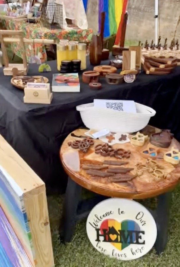 Display table with various wooden crafts and baked goods, including cookie shapes. A sign reads 'Welcome to Our Home - Love Lives Here,' featuring a rainbow and heart design.