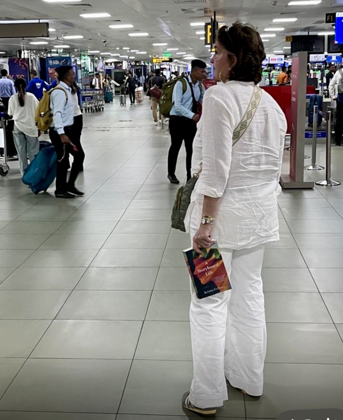 A woman in white pants and a white blazer, carrying a book titled 'Storyboard Life,' stands in an airport terminal with travelers walking around and luggage visible.