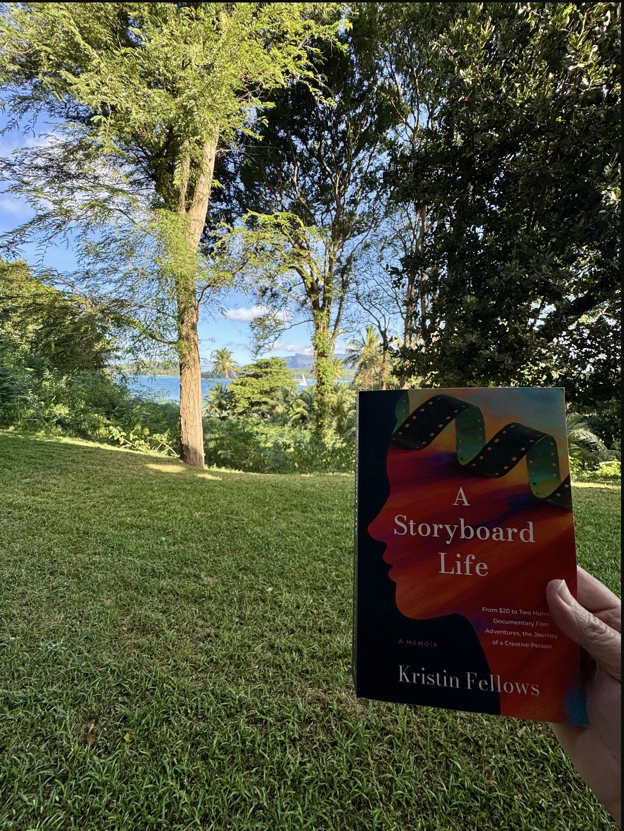 A person holding a book titled 'A Storyboard Life' by Kristin Fellows outdoors in a lush, green, tropical setting with trees, grass, and a view of the ocean and sailboats in the distance.