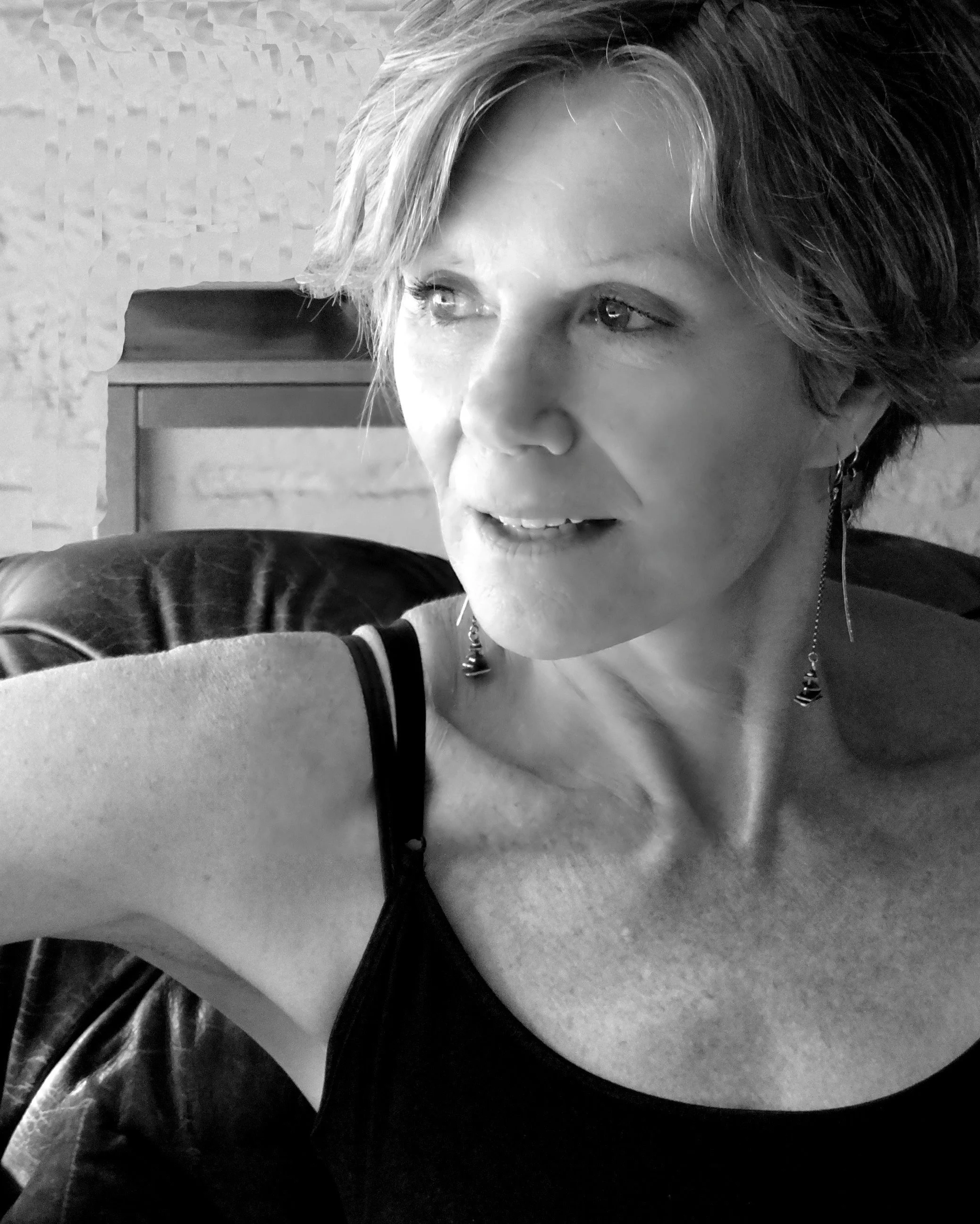 A close-up black-and-white photo of a woman with short, wavy hair, wearing earrings and a black top, looking slightly to the side.