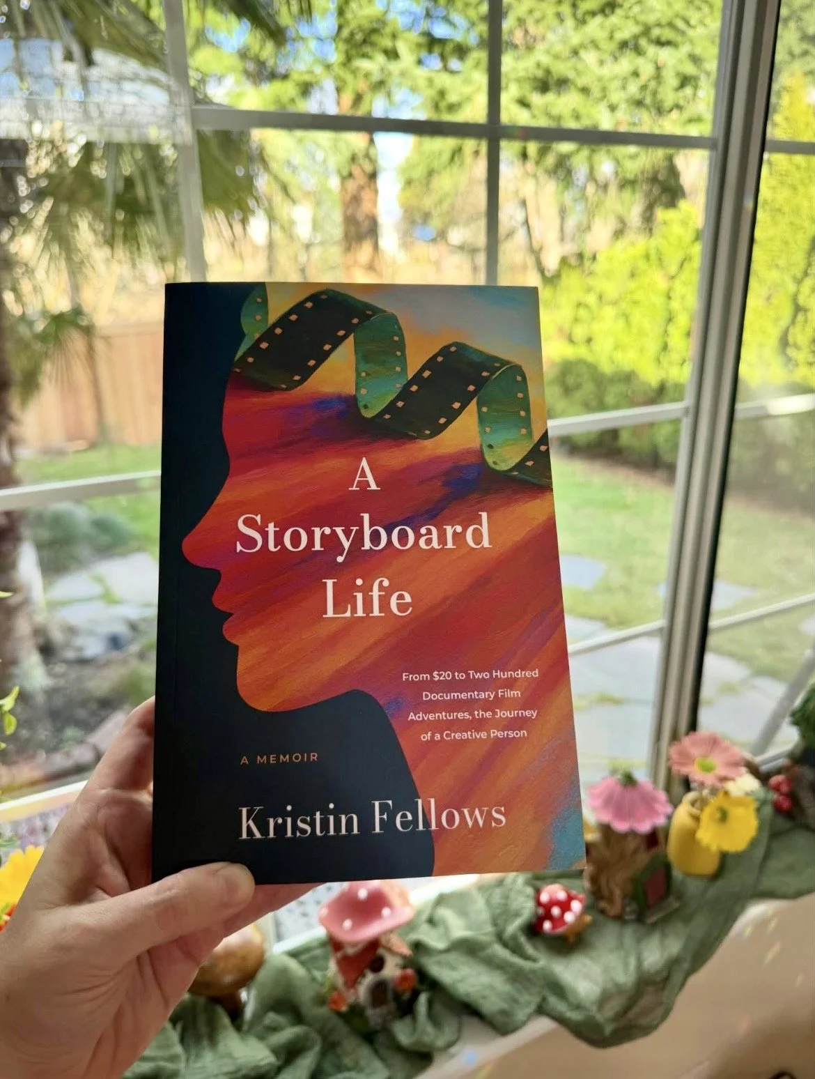 A hand holds a book titled 'A Storyboard Life' by Kristin Fellows, with a colorful cover depicting a silhouette of a person's face and a film strip. The background shows a window with trees and greenery outside, and a decorative scene with small mushroom and flower ornaments inside.