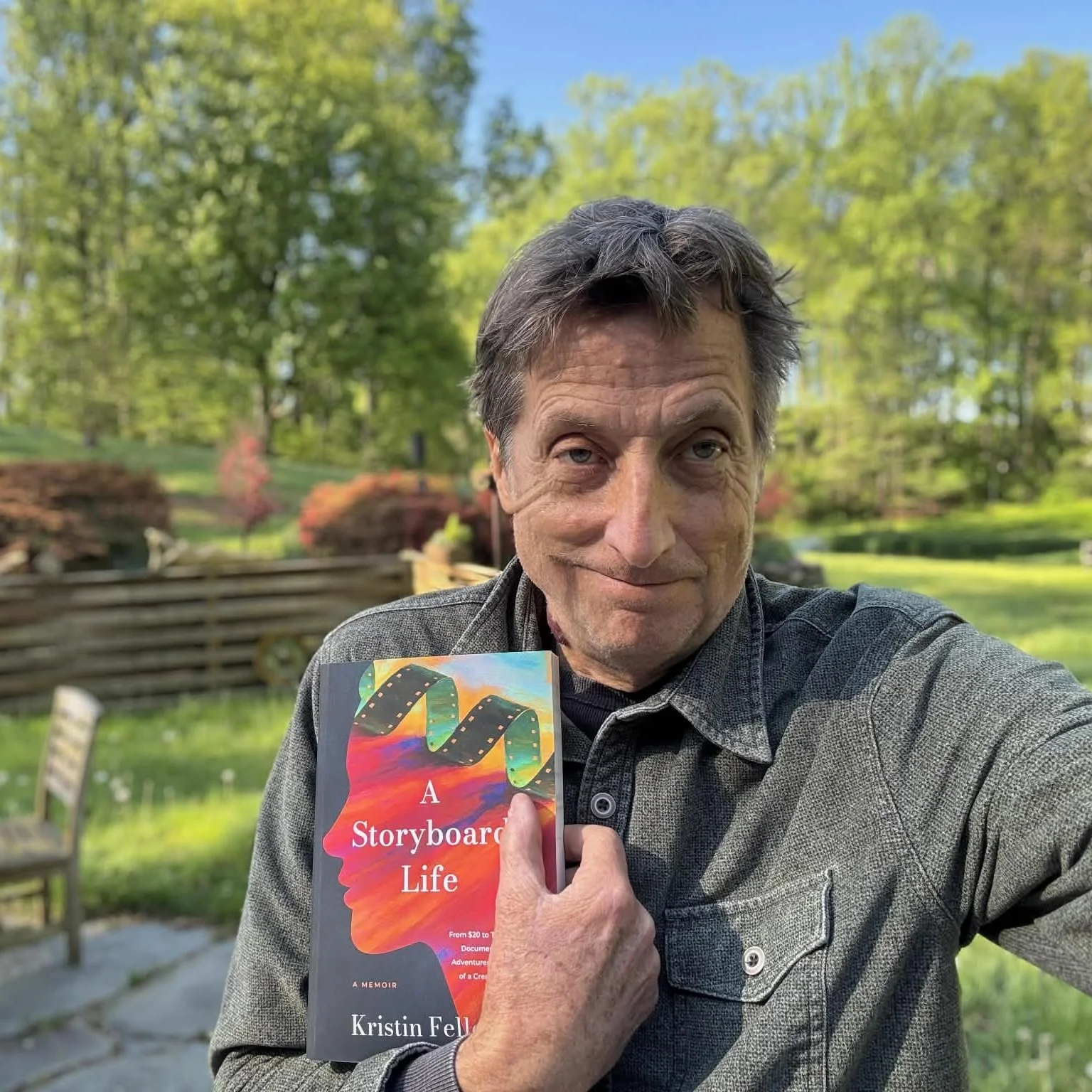 A middle-aged man with short, tousled hair, smiling slightly, holding a book titled "A Storyboard Life" by Kristin Fels in an outdoor setting with green trees and a wooden fence in the background.
