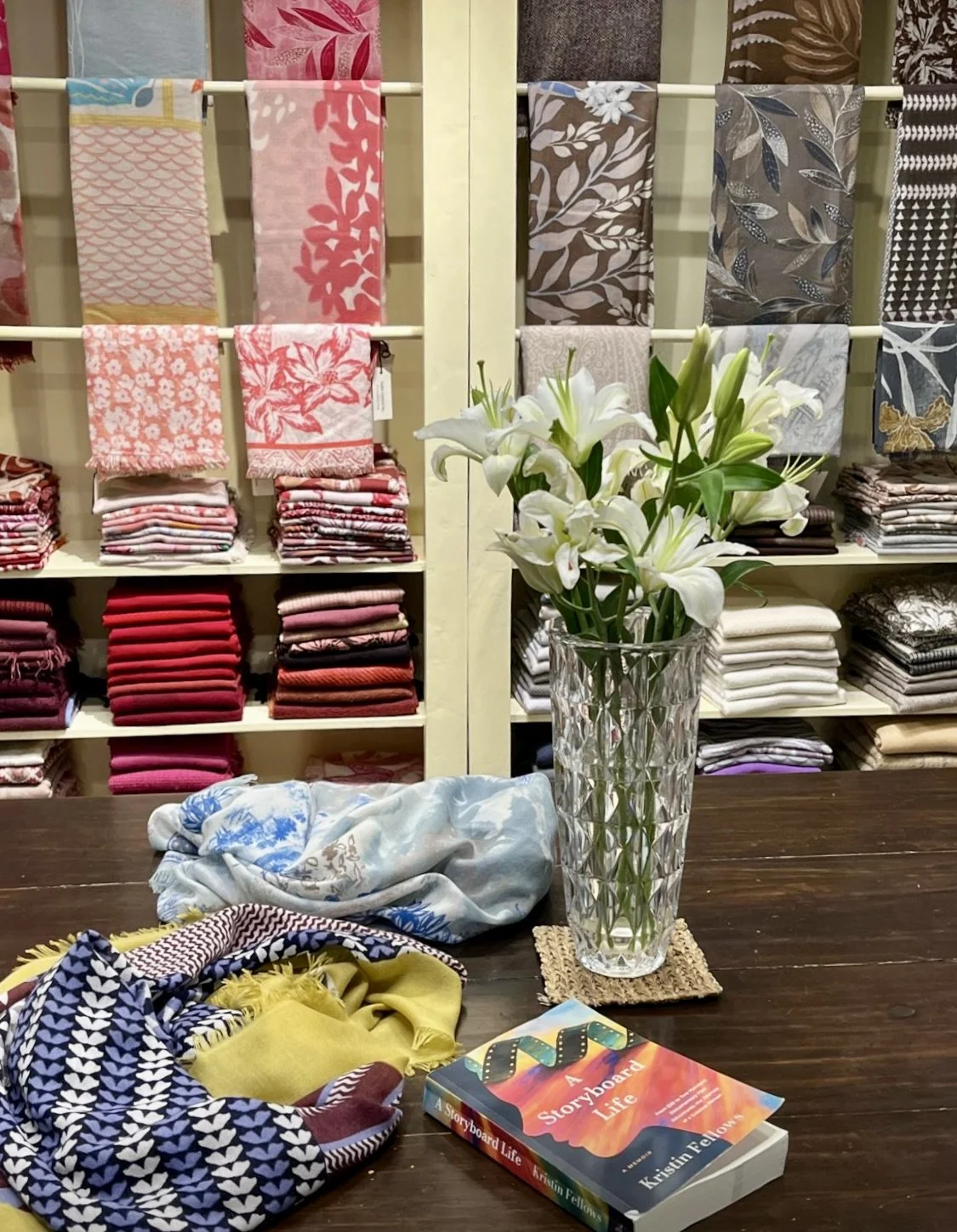 A dark wooden table with a bouquet of white lilies in a glass vase, a colorful book titled "Storyboard Life" by Kristin Felos, and some fabric swatches. In the background, there are shelves stocked with folded fashionable fabrics and textiles in various colors and patterns.