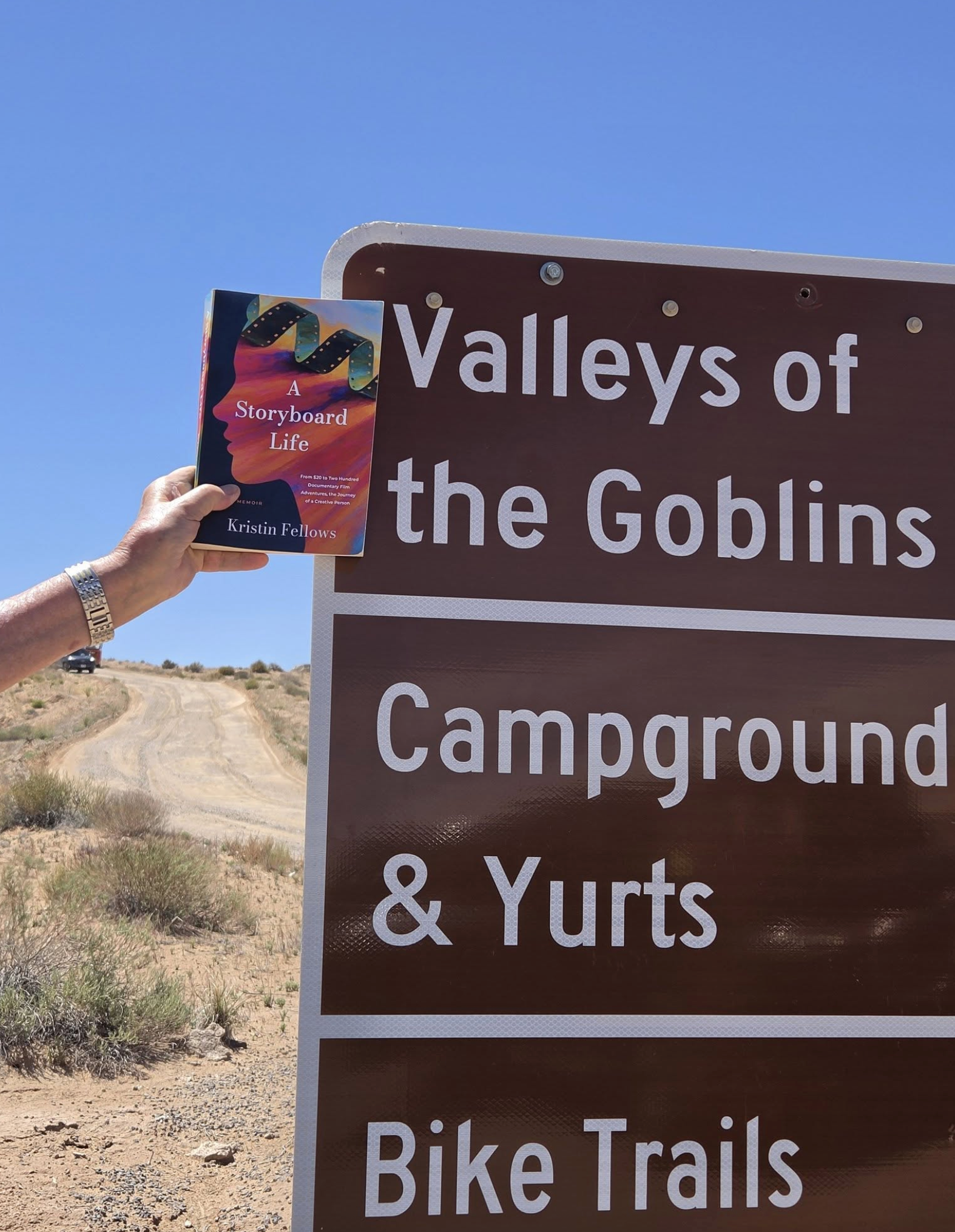 A hand holding a brochure in front of a sign that reads "Valleys of the Goblins, Campground, & Yurts" on a dirt trail in a dry, open landscape under a clear blue sky.