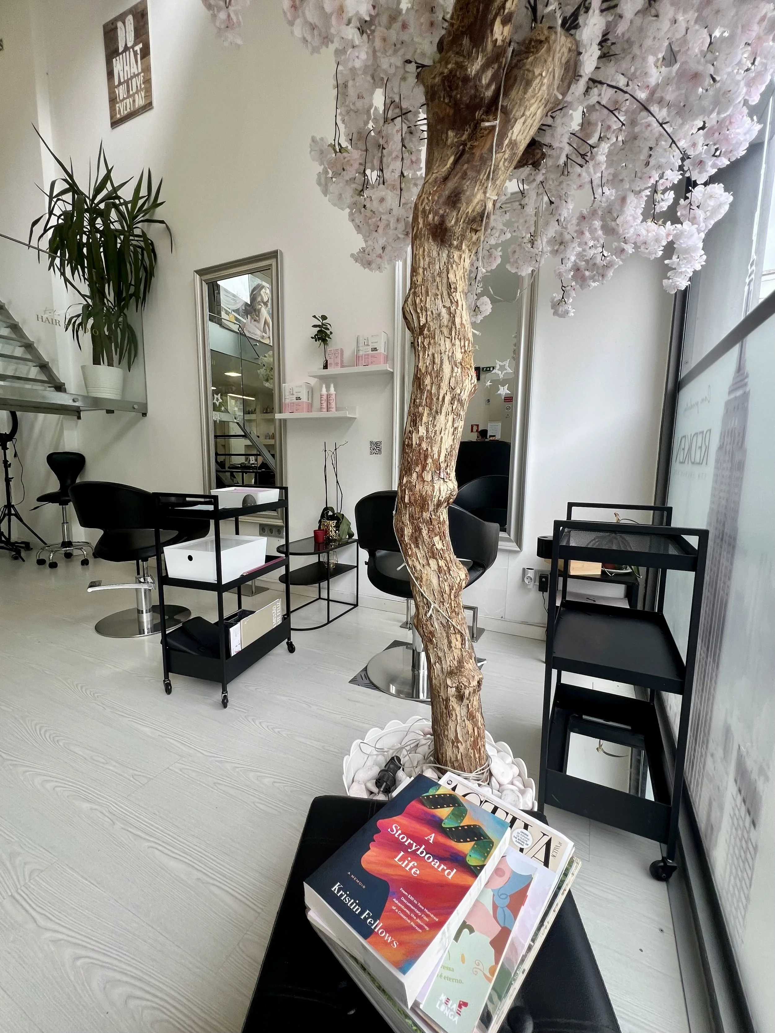 Indoor setting with a prominent artificial cherry blossom tree in a white pot, surrounded by black chairs, a small table with books, a mirror, and various office supplies and decorations, including a plant and framed wall art.