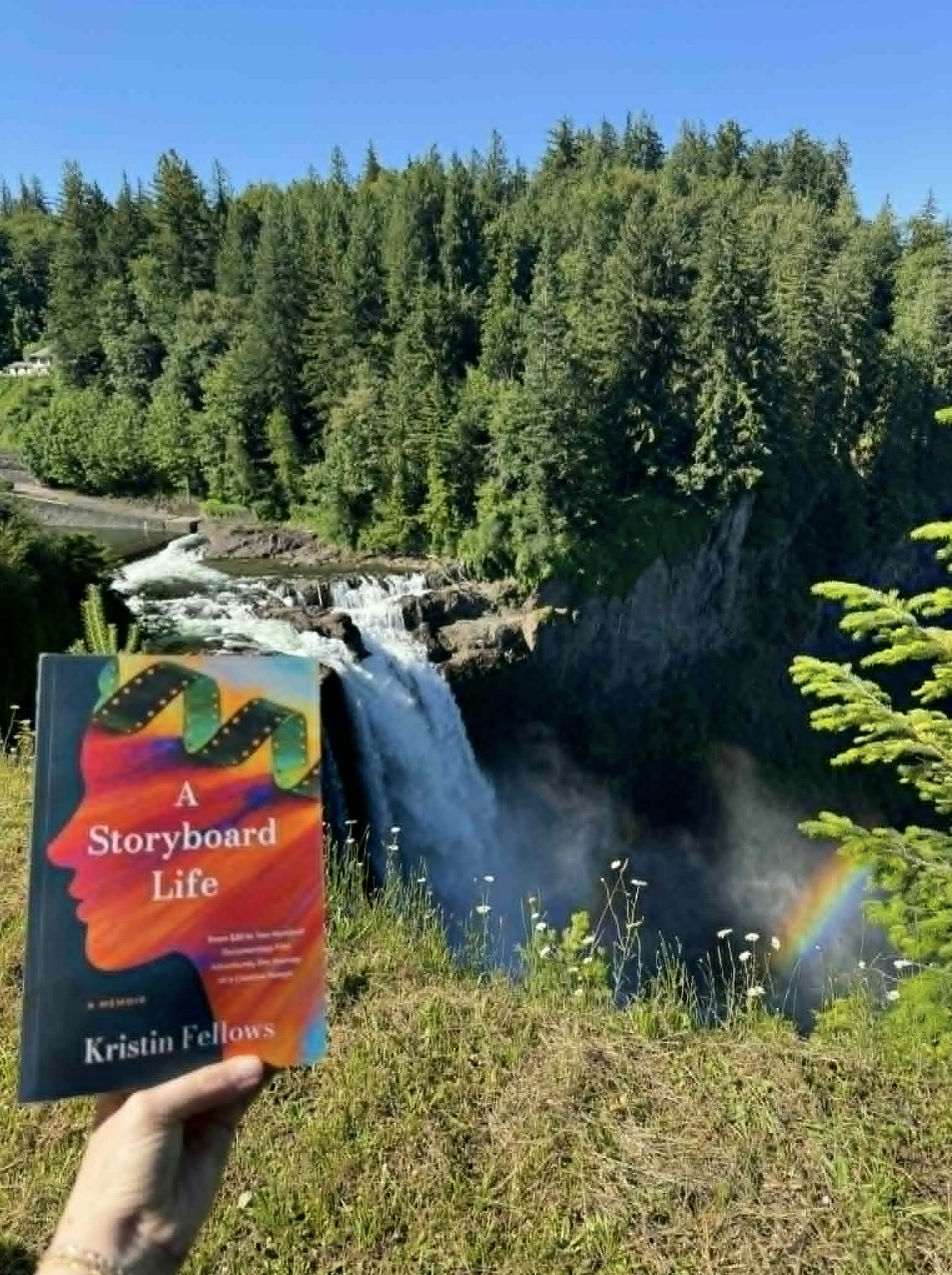 A person holding a book titled "A Storyboard Life" by Kristin Fellows in front of a waterfall, with a rainbow visible near the bottom right corner of the image. The background features a lush, green forest and a bright blue sky.