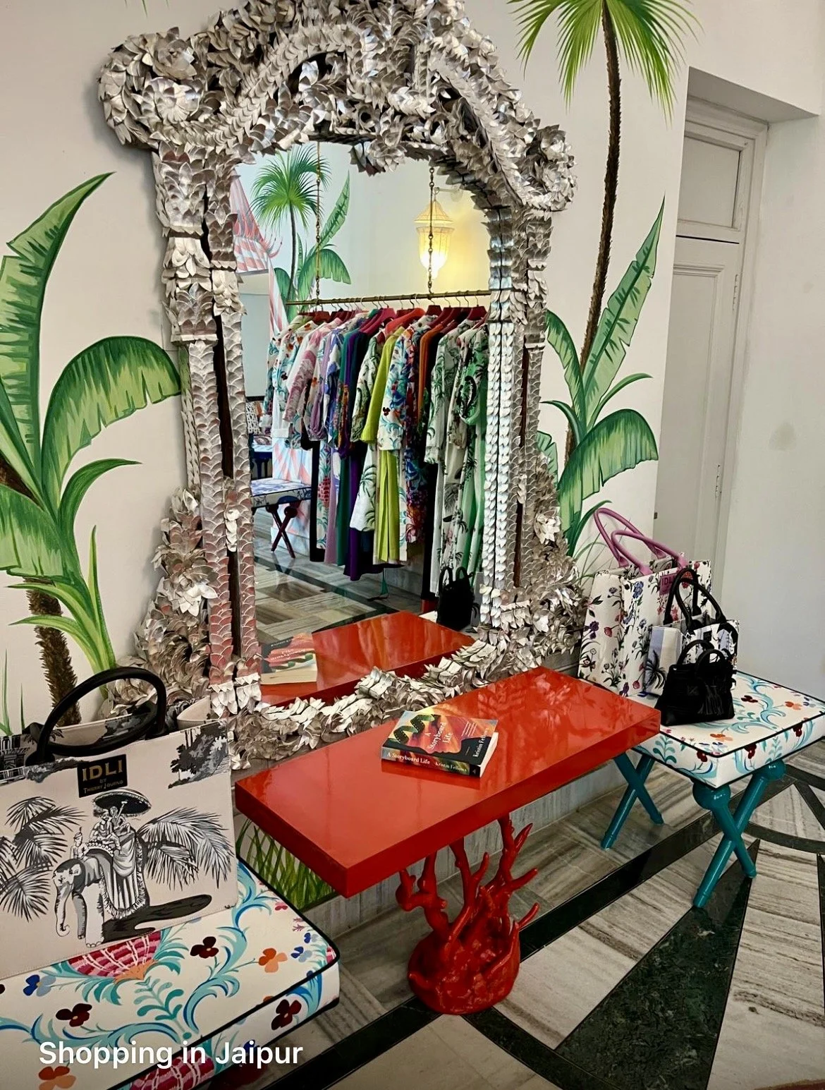 A mirror with a decorative, leafy frame reflects a clothing rack with vibrant, tropical patterned dresses, and a tropical-themed mural with palm trees in the background. In front of the mirror, there are colorful tables and chairs, with shopping bags and a book on the tables, indicating a boutique shop in Jaipur.