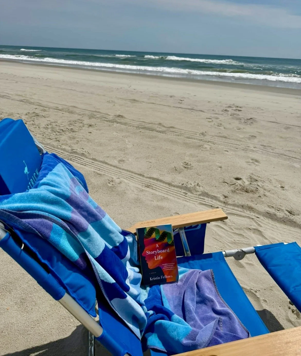 A beach scene with an empty blue lounge chair and a colorful book titled 'A Storyboard Life' beside it. The sandy beach leads to the ocean with small waves, under a mostly cloudy sky.