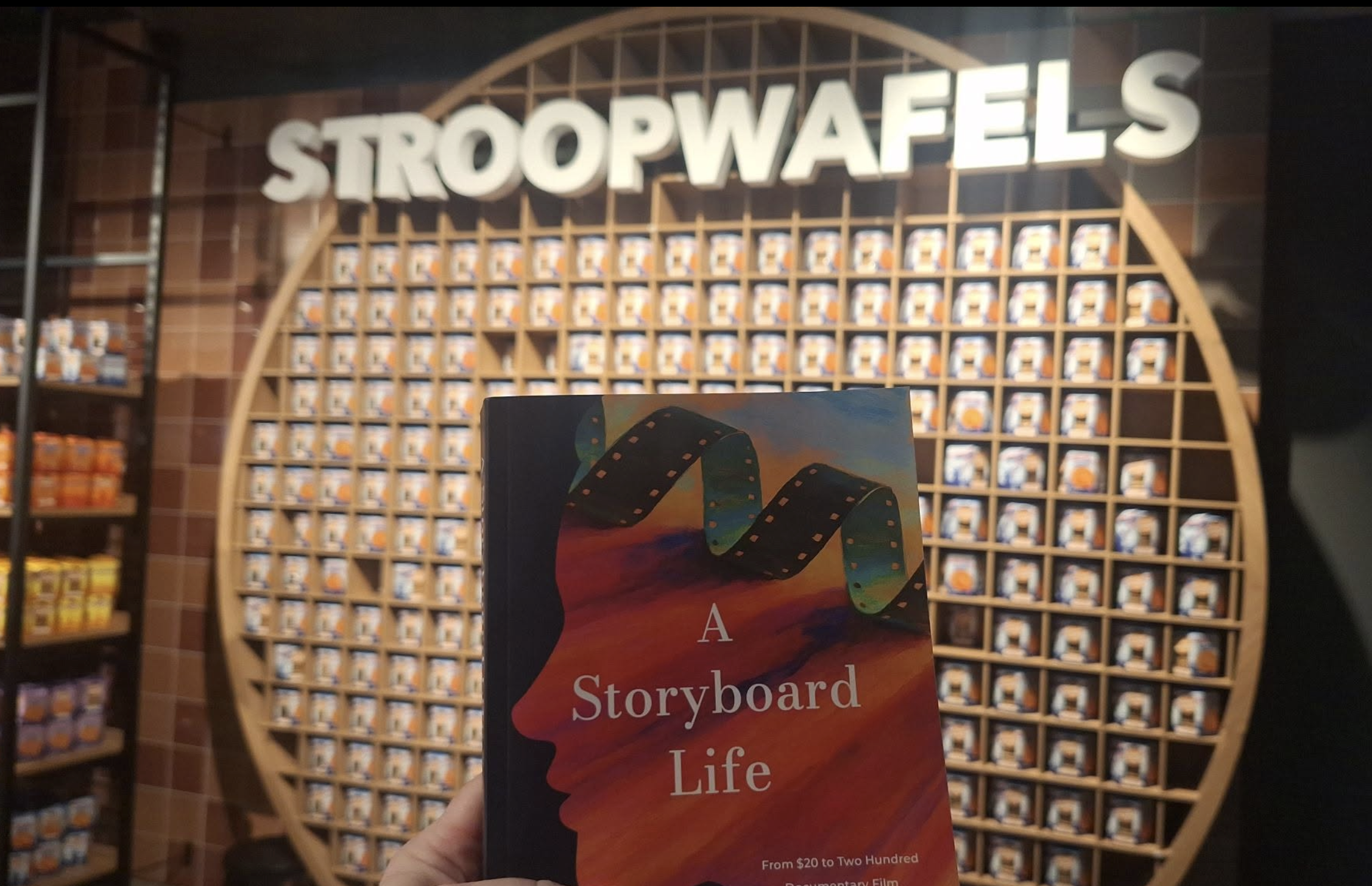A person holding a colorful brochure with a film strip graphic and text that reads 'A Storyboard of Life' in front of a display wall filled with small boxes, and a large sign that says 'STROOPWAFELS' in the background.