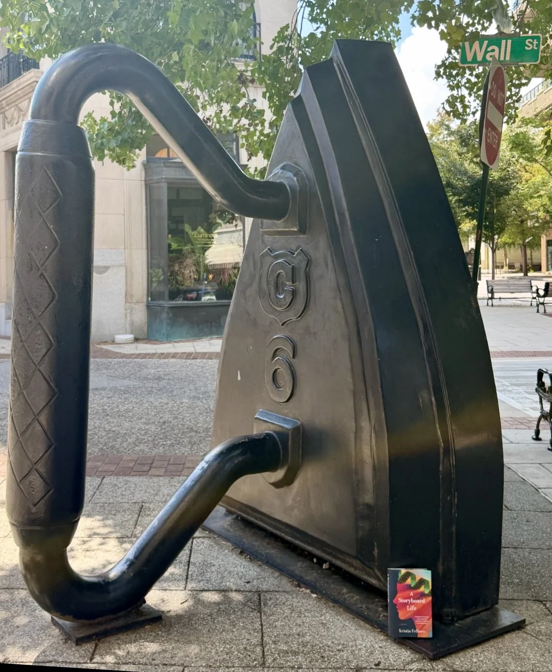 A large, black, metal sculpture of a typewriter placed outdoors on a city sidewalk with trees and store windows in the background. There are street signs for Wall Street and a stop sign visible.