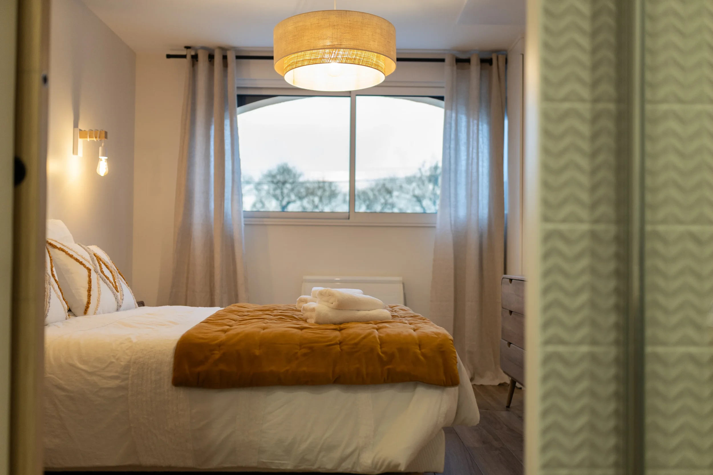 Chambre d'hôtel avec lit fait, deux serviettes pliées, grande fenêtre avec vue sur des arbres, rideaux clairsemés, lampes de chevet, plafond avec luminaire ronde