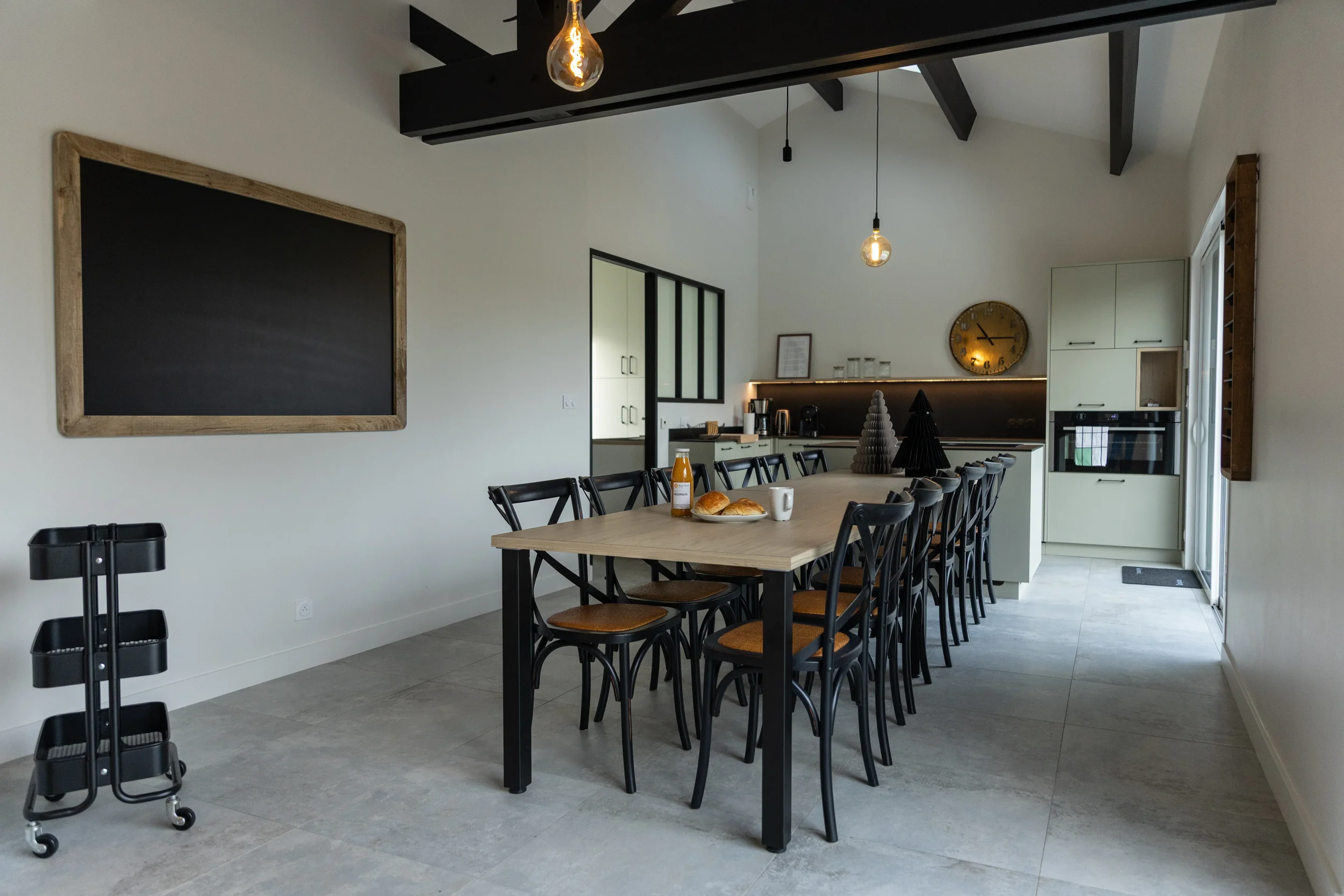 Salle à manger moderne avec grande table en bois, plusieurs chaises noires avec assises marron, décor minimaliste, cuisine en arrière-plan, horloge ronde dorée, lampes suspendues
