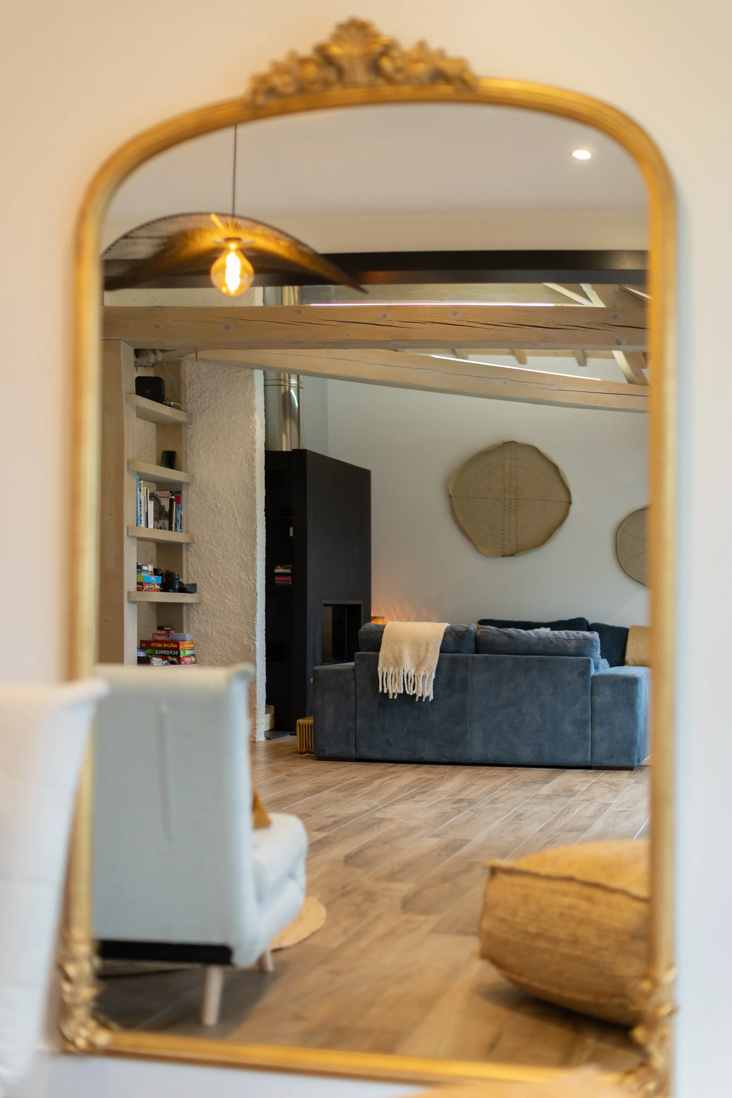 Un salon moderne avec un canapé bleu, une étagère remplie de livres, un mur décoratif rond, et un plafond en bois, vu dans un miroir avec un cadre doré.