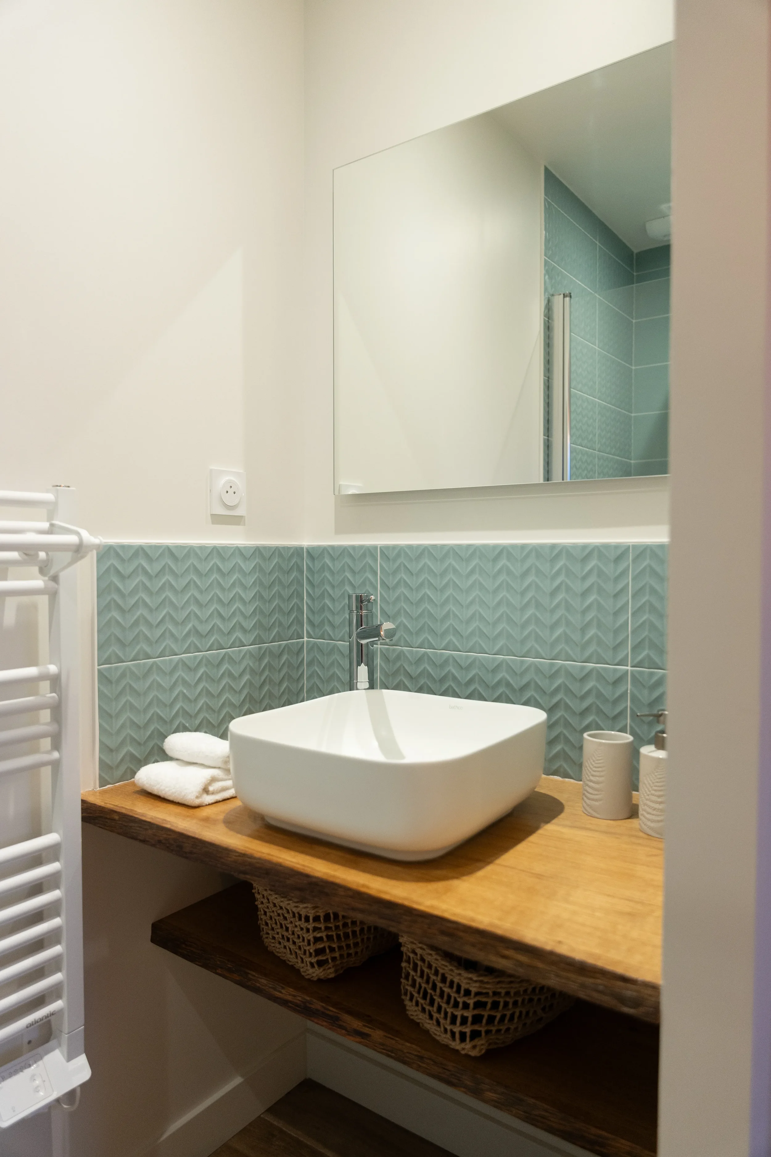 Un lavabo moderne sur une planche en bois, avec un miroir rectangulaire au mur, derrière une salle de bain avec carrelage vert et des accessoires en céramique, dans une salle de bain élégante.