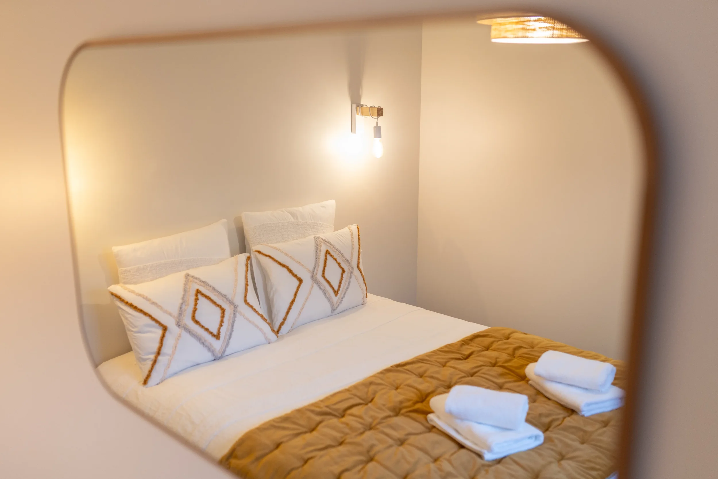 Lit double dans une chambre, vue à travers un miroir, avec oreillers blanche à motifs dorés, couverture beige, serviettes blanches pliées sur le lit, éclairage chaud.