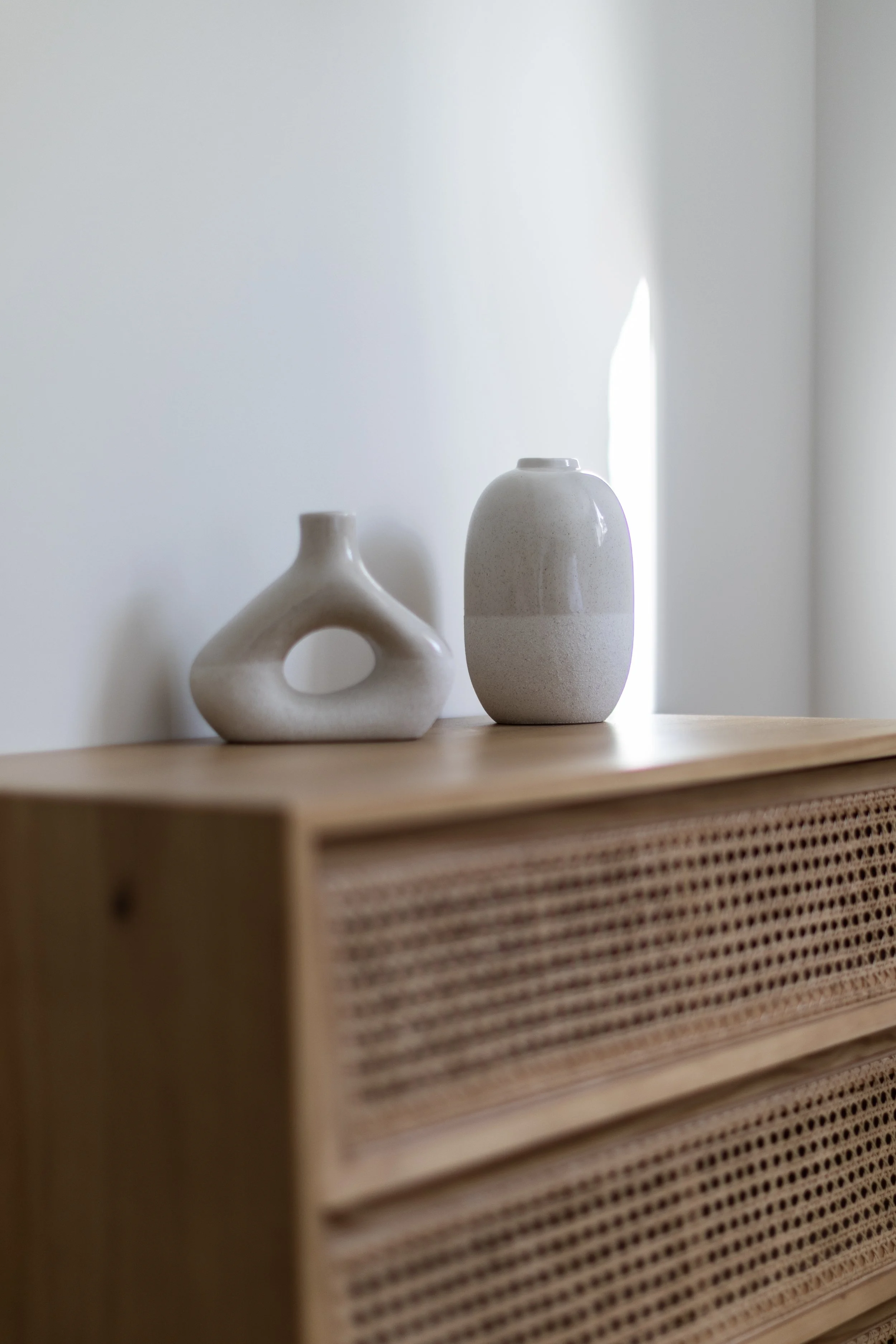 Deux vases en céramique, un avec une forme géométrique ouverte et l'autre cylindrique, posés sur un meuble en bois avec un motif en rotin.