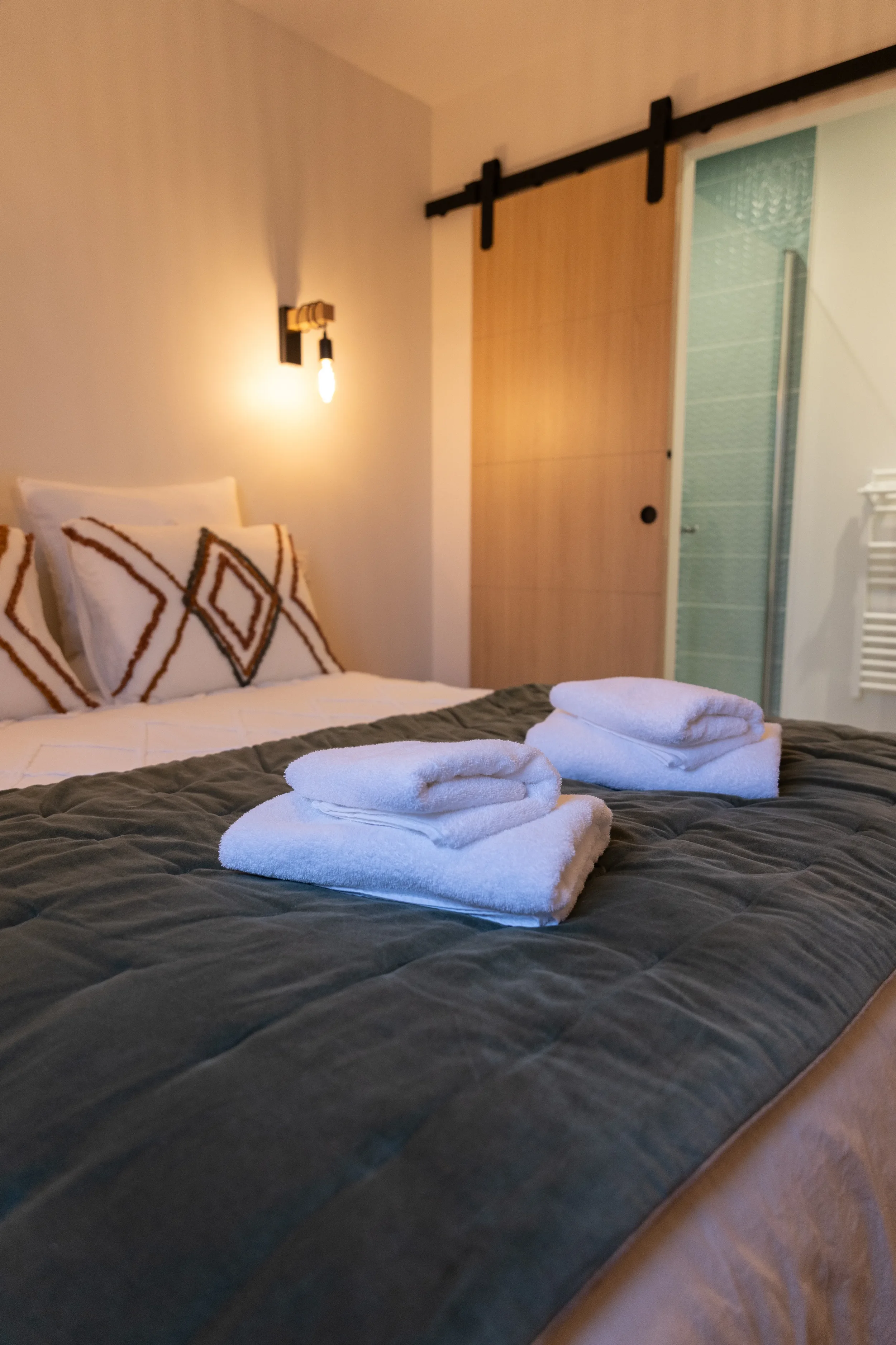 Lits avec des serviettes blanches pliées sur la couverture. Nuit, ambiance chaleureuse avec éclairage doux, porte coulissante en bois, murs clairs et décoration moderne.