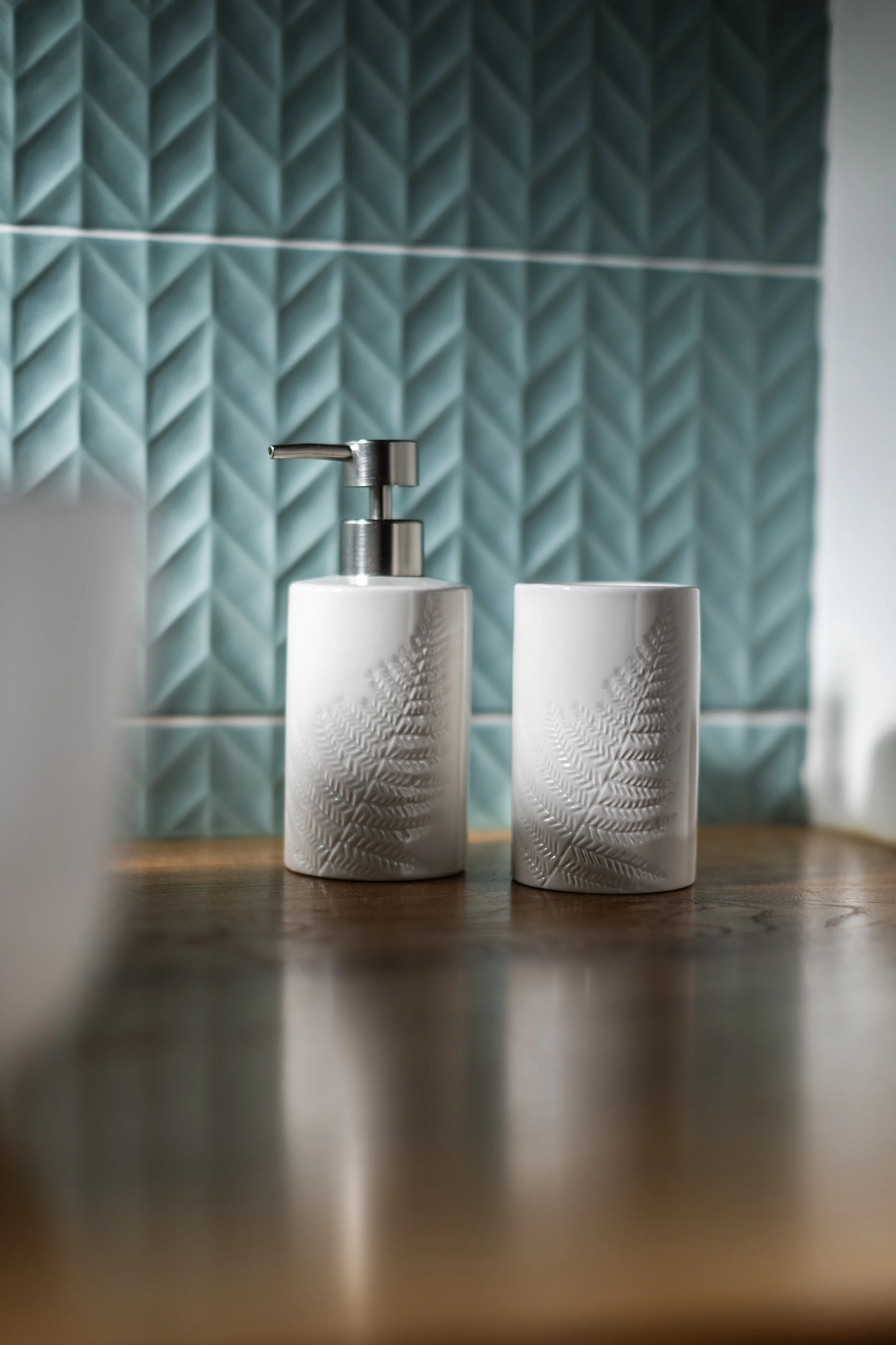 Distributeurs de savon et de lotion en céramique blanche avec motif de fougère, sur un plan de travail en bois, avec un mur en carrelage bleu en arrière-plan.