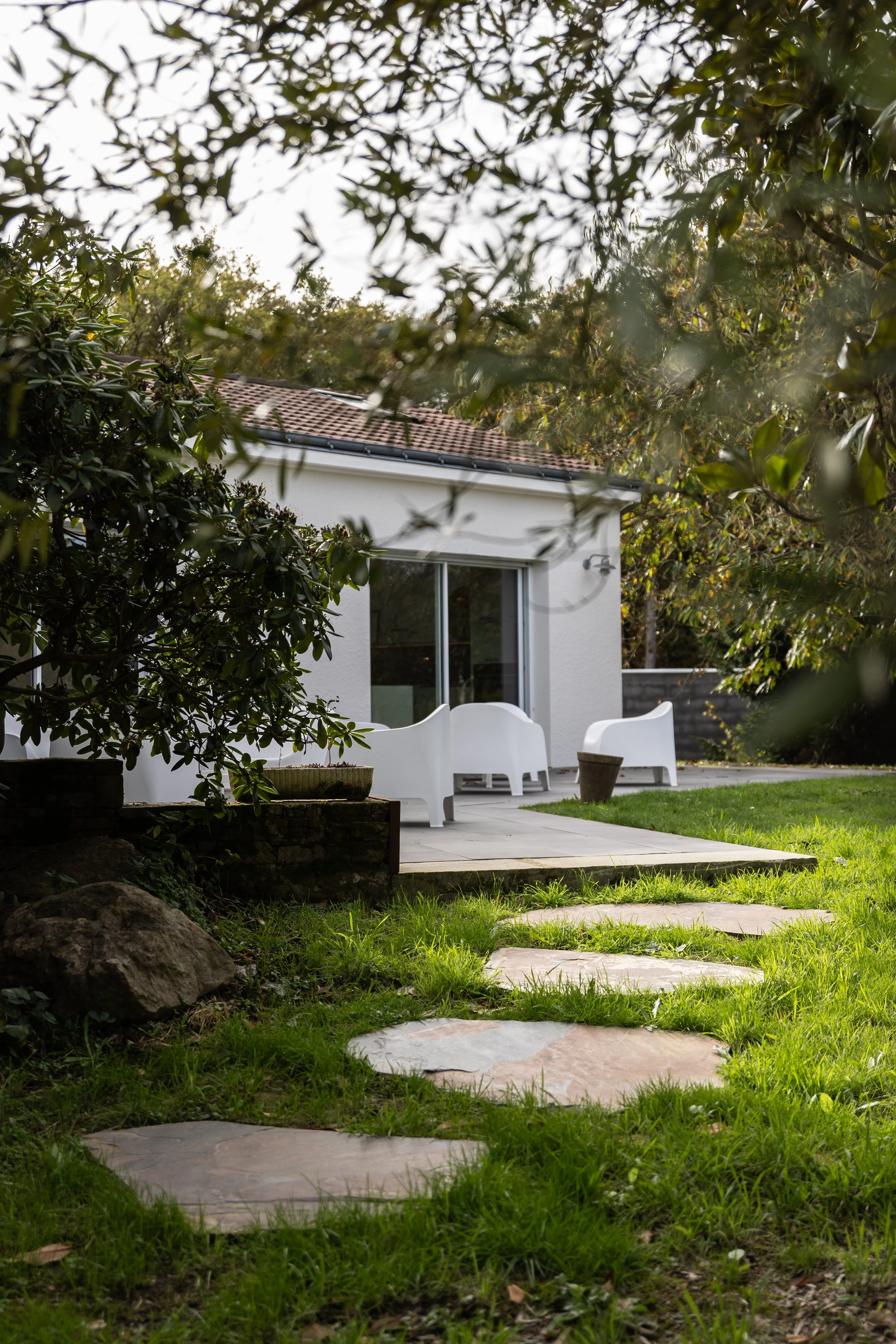 Une maison moderne avec une terrasse, entourée d'arbres et de gazon vert, avec des pierres plates formant un chemin vers la terrasse.