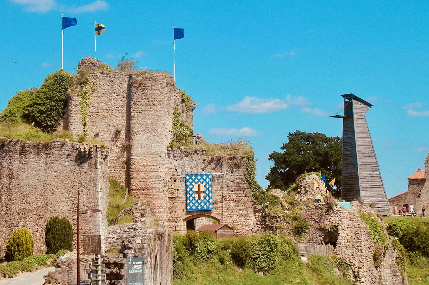 Restes du château de TIFFAUGE médiéval en pierre avec drapeaux, une bannière bleue avec une croix, un pont en pierre et une tour en bois, sous un ciel bleu avec quelques nuages.