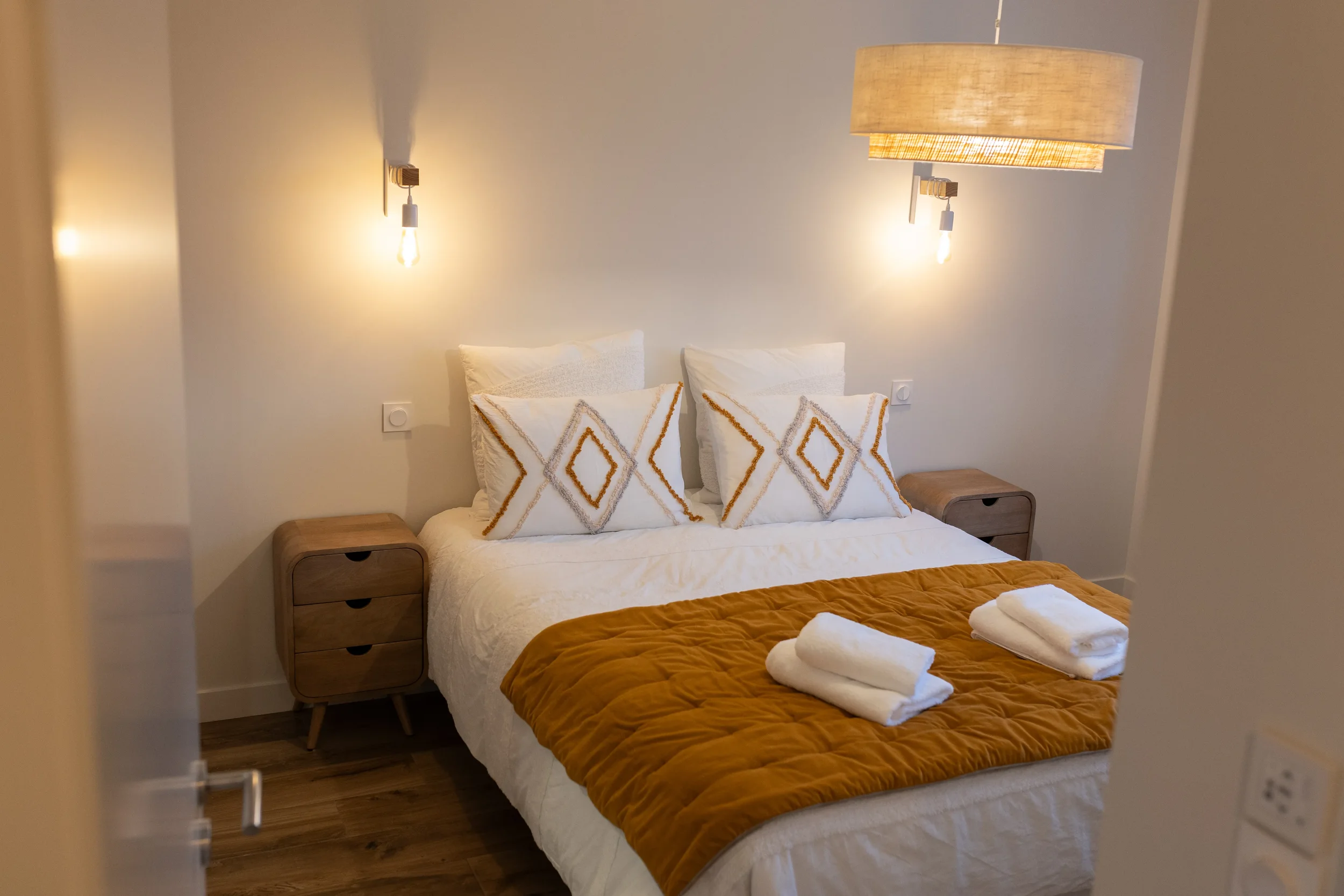 Chambre avec lit double, deux petites tables de nuit en bois, deux lampes murales, coussins décoratifs, couverture marron, serviettes blanches sur le lit, sol en bois clair.