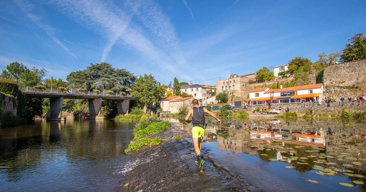 Un homme courant sur un chemin le long d'une rivière dans un village avec des bâtiments colorés, un pont et des arbres, par une journée ensoleillée.