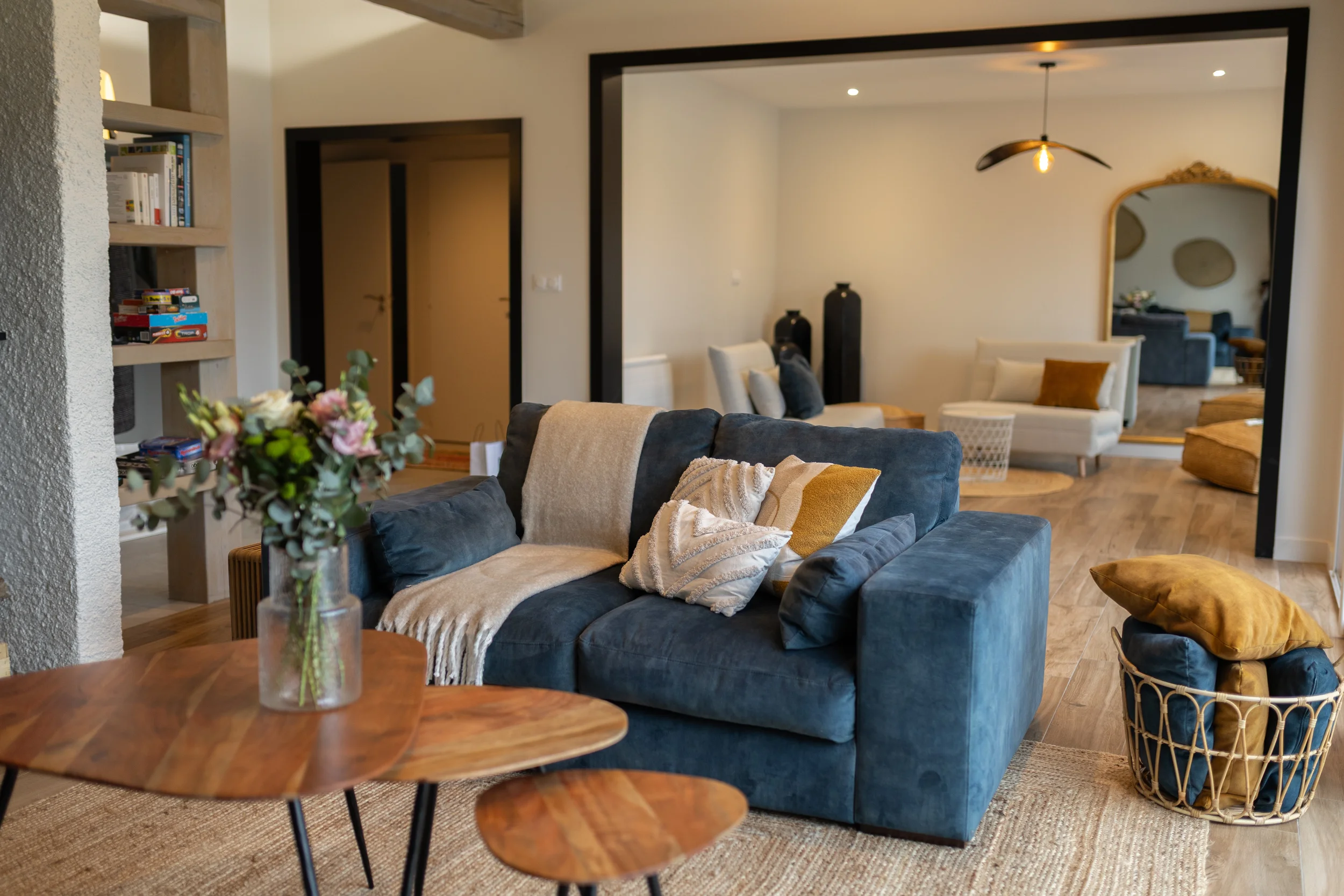 Salon moderne avec un canapé bleu, coussins, un tapis beige, une table en bois avec des fleurs, et un espace avec d'autres canapés et un miroir doré.