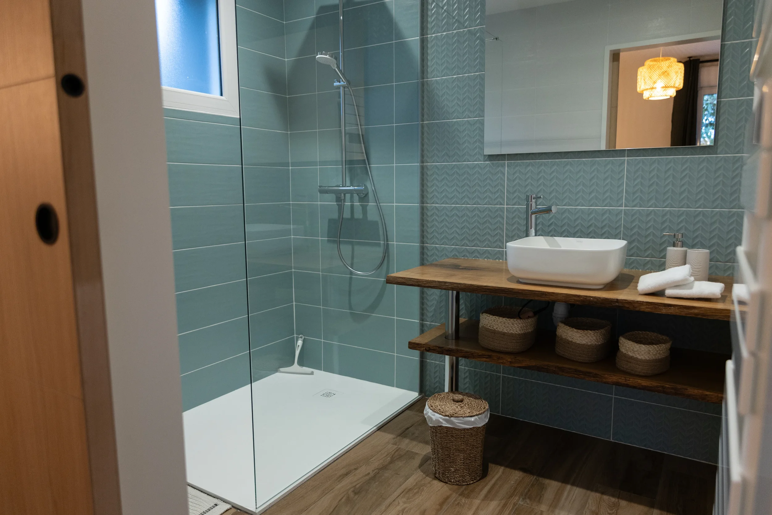 Salle de bain avec douche, lavabo sur un meuble en bois, miroir, serviettes blanches, paniers en rotin, sol en bois.
