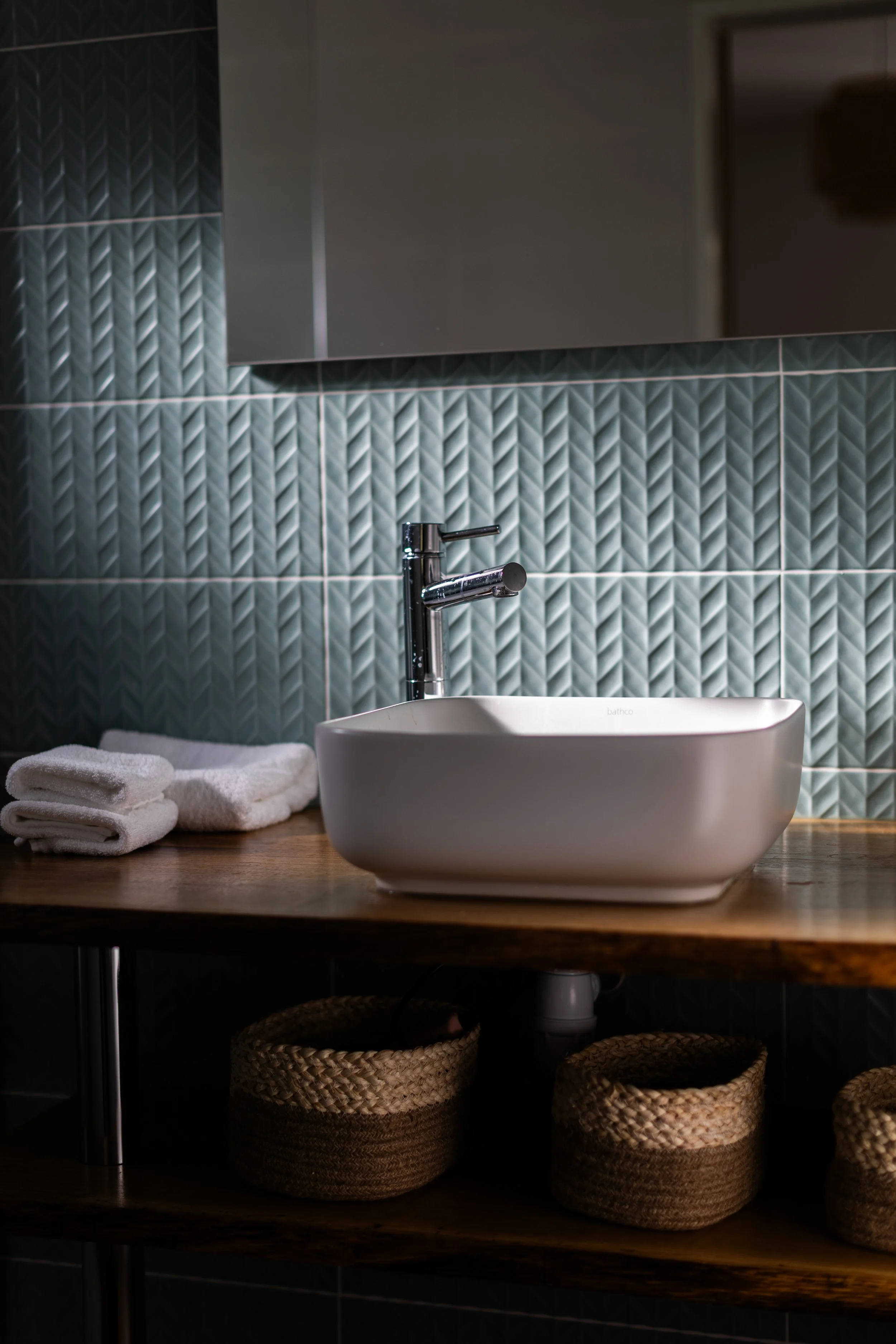Lavabo blanc avec robinet, serviettes blanches pliées à gauche, paniers en osier sous le plan de travail en bois, mur en carrelage vert avec motif géométrique, miroir au-dessus du lavabo.