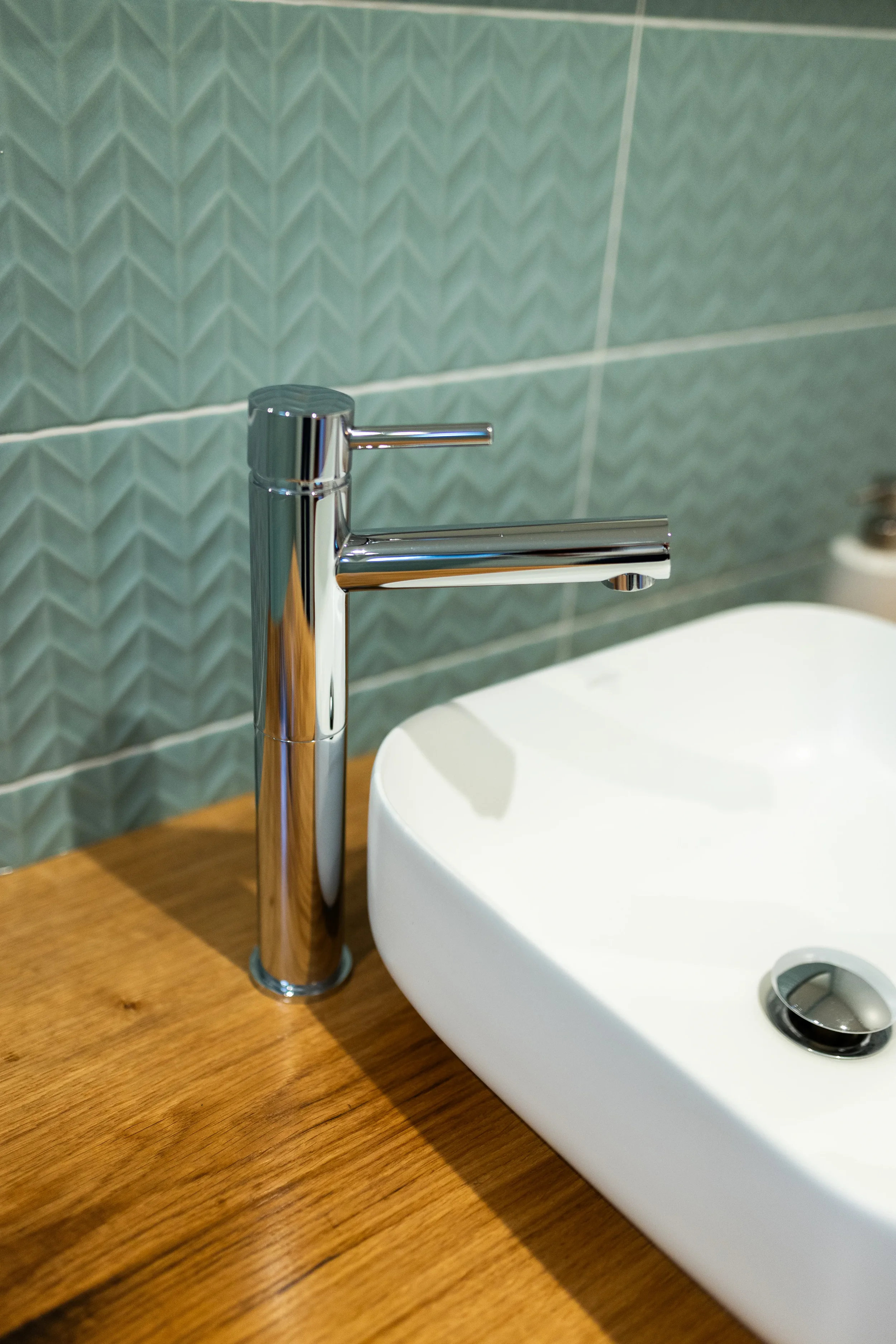 Un robinet en chrome à côté d'un évier blanc posé sur un comptoir en bois, avec un mur en carreaux verticaux derrière.