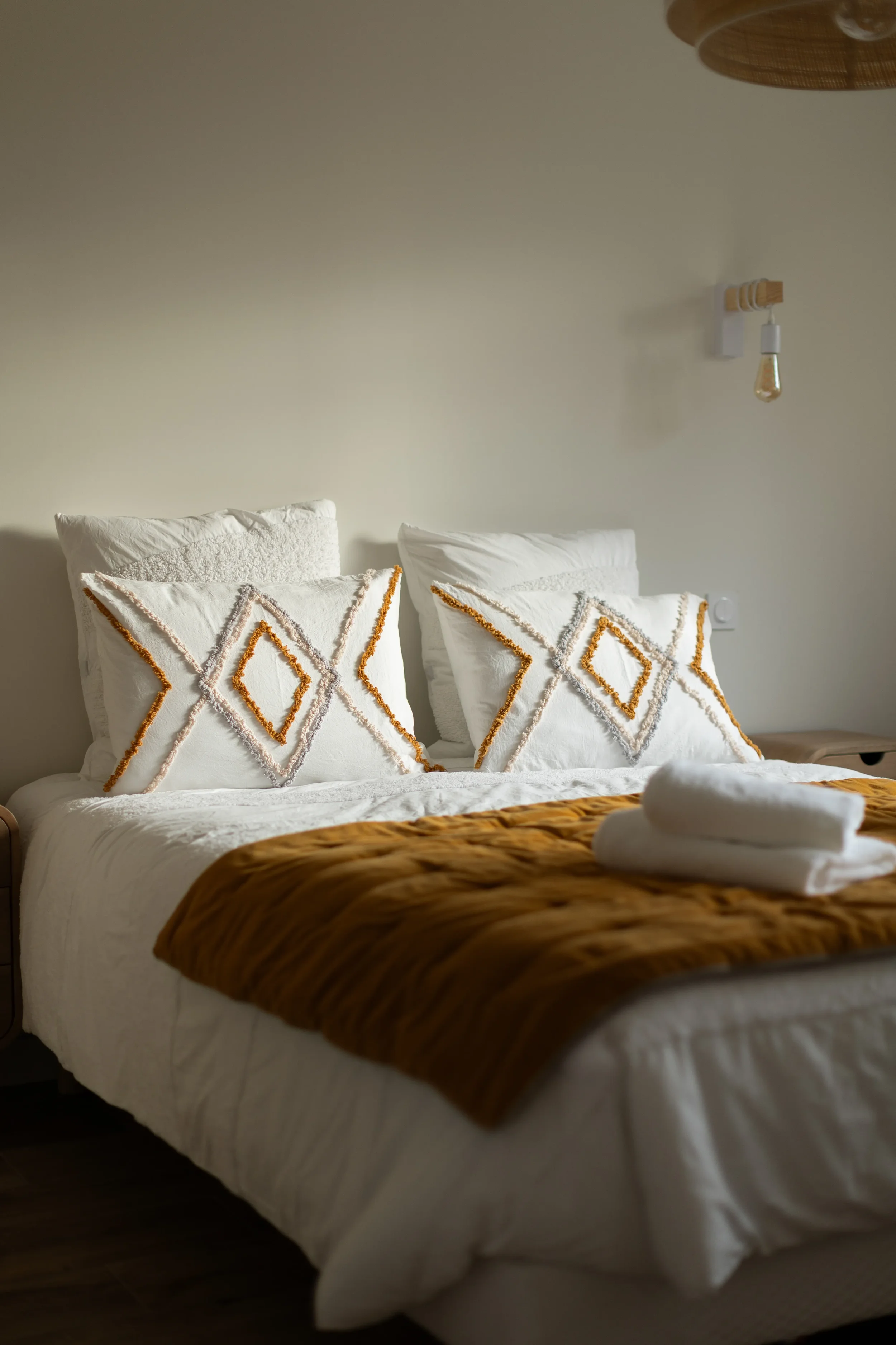Lits doubles avec deux oreillers blancs ornés de motifs géométriques en orange et gris, couverture marron, serviettes blanches pliées sur le lit, mur beige avec une lampe à ampoule apparente au mur.