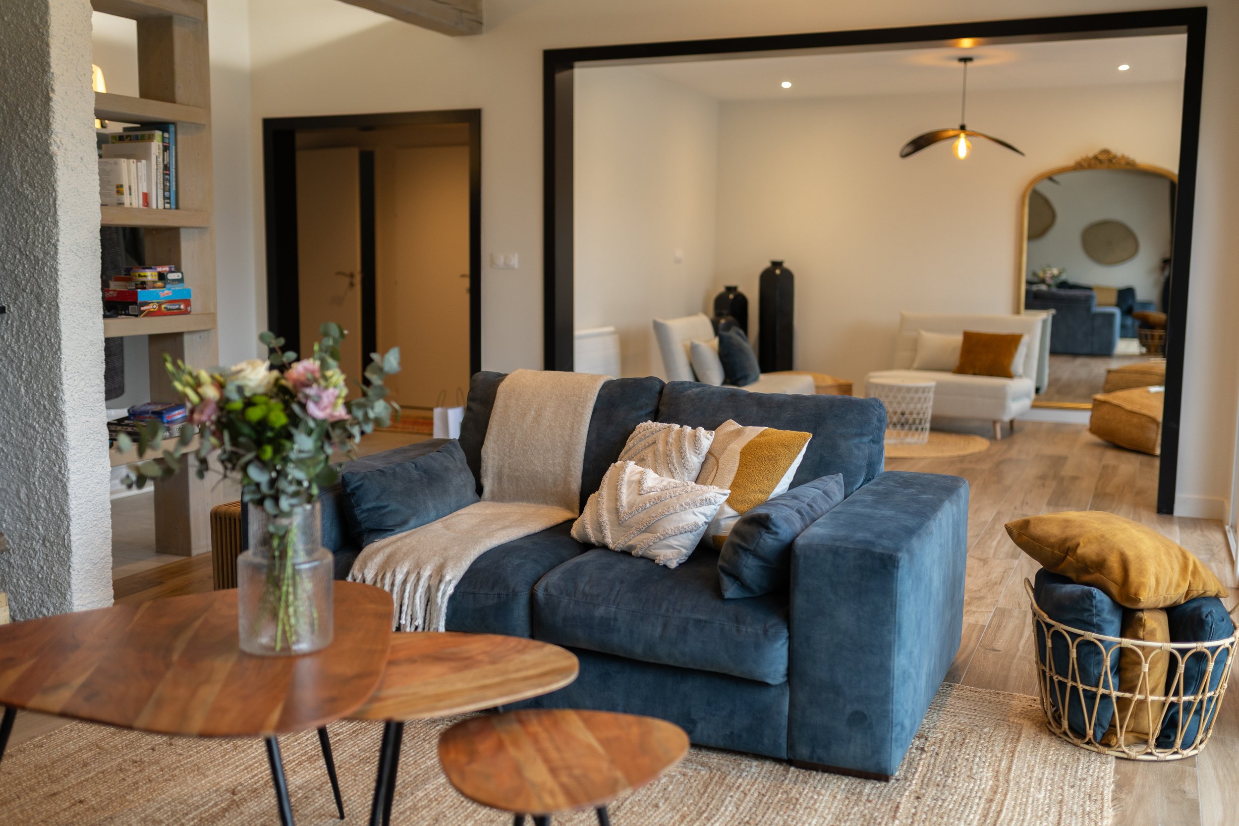 Salon avec un canapé bleu, une table en bois avec un vase de fleurs, et un fond d'espace salon séparé par une ouverture noire.