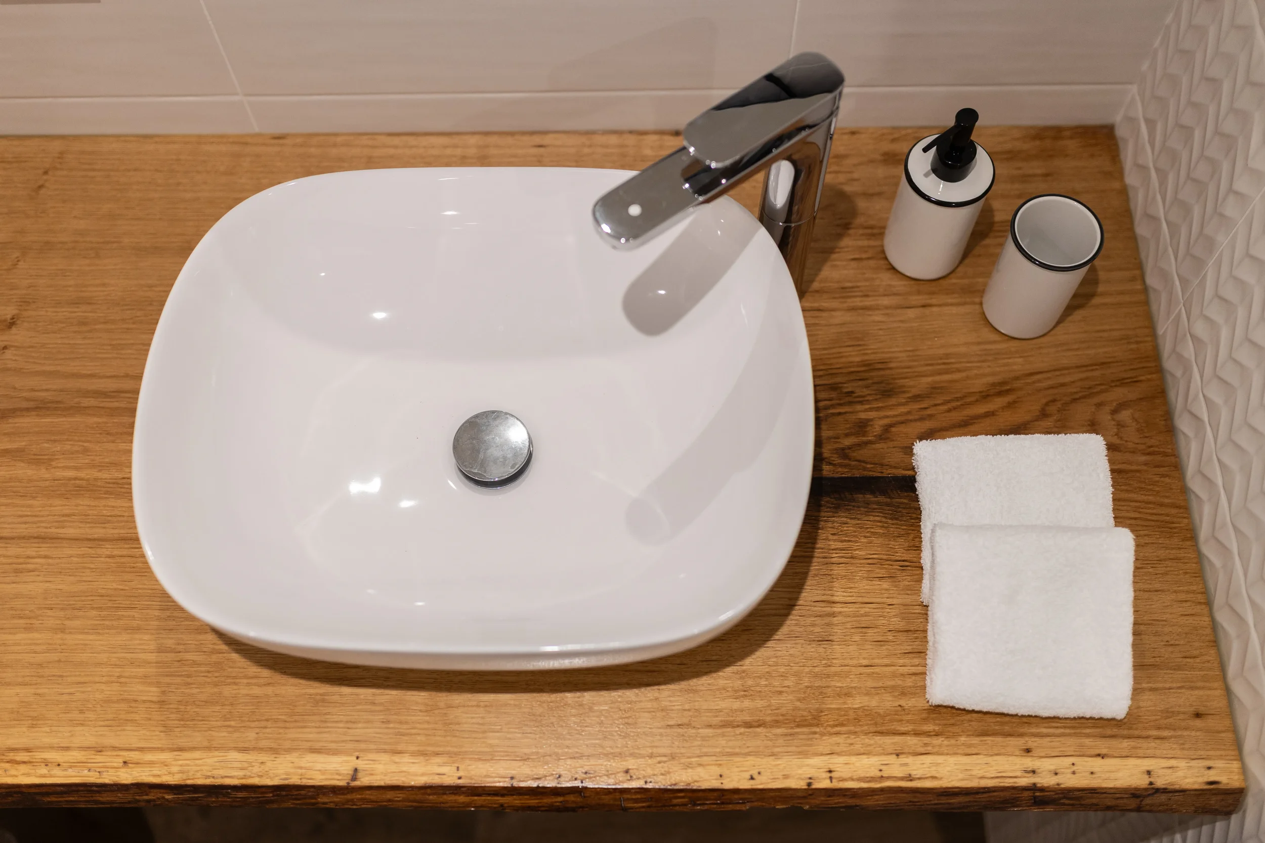 Lavabo blanc carré sur plan en bois, avec robinet en chrome, savon en bouteille avec pompe, mug blanc avec finition noire, deux serviettes blanches pliées