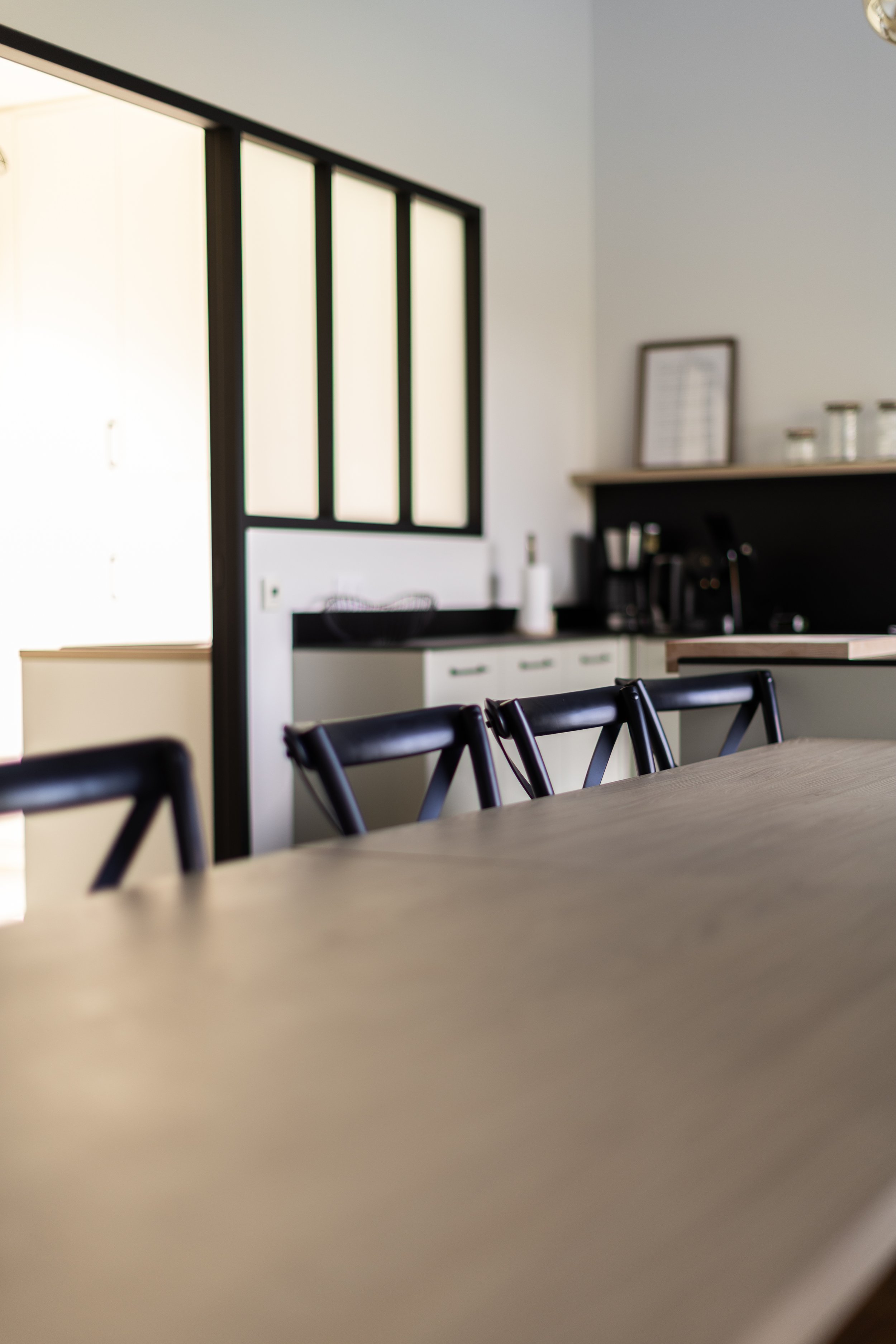 Une table en bois avec plusieurs chaises en bois noires dans une cuisine moderne, visible partiellement avec des armoires blanches et des éléments noirs, et une fenêtre en fond lumineux.