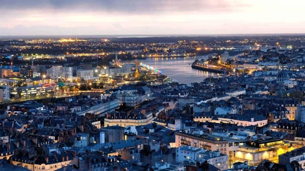 Vue aérienne de la ville de Nantes éclairée la nuit avec un fleuve au centre et des bâtiments modernes et anciens.